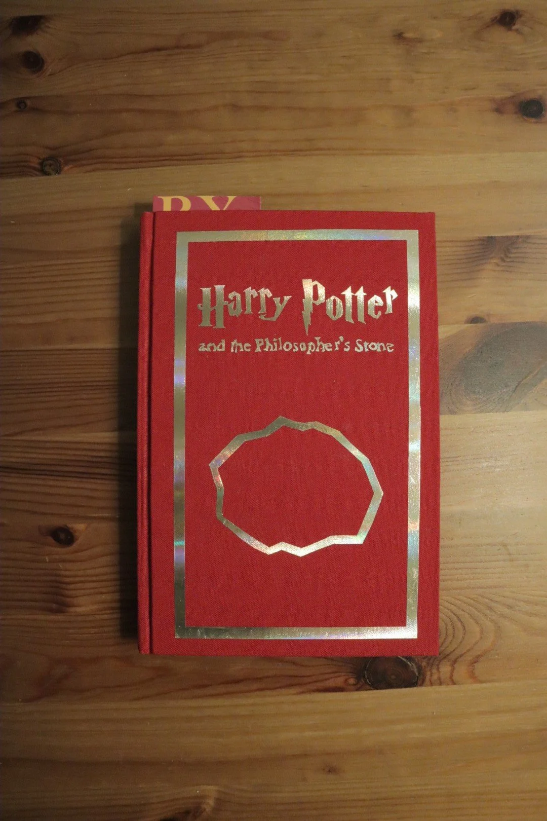 A red hardcover book titled 'Harry Potter and the Philosopher's Stone' with a white and gold border and a gold outlined circle on the cover, placed on a wooden surface.