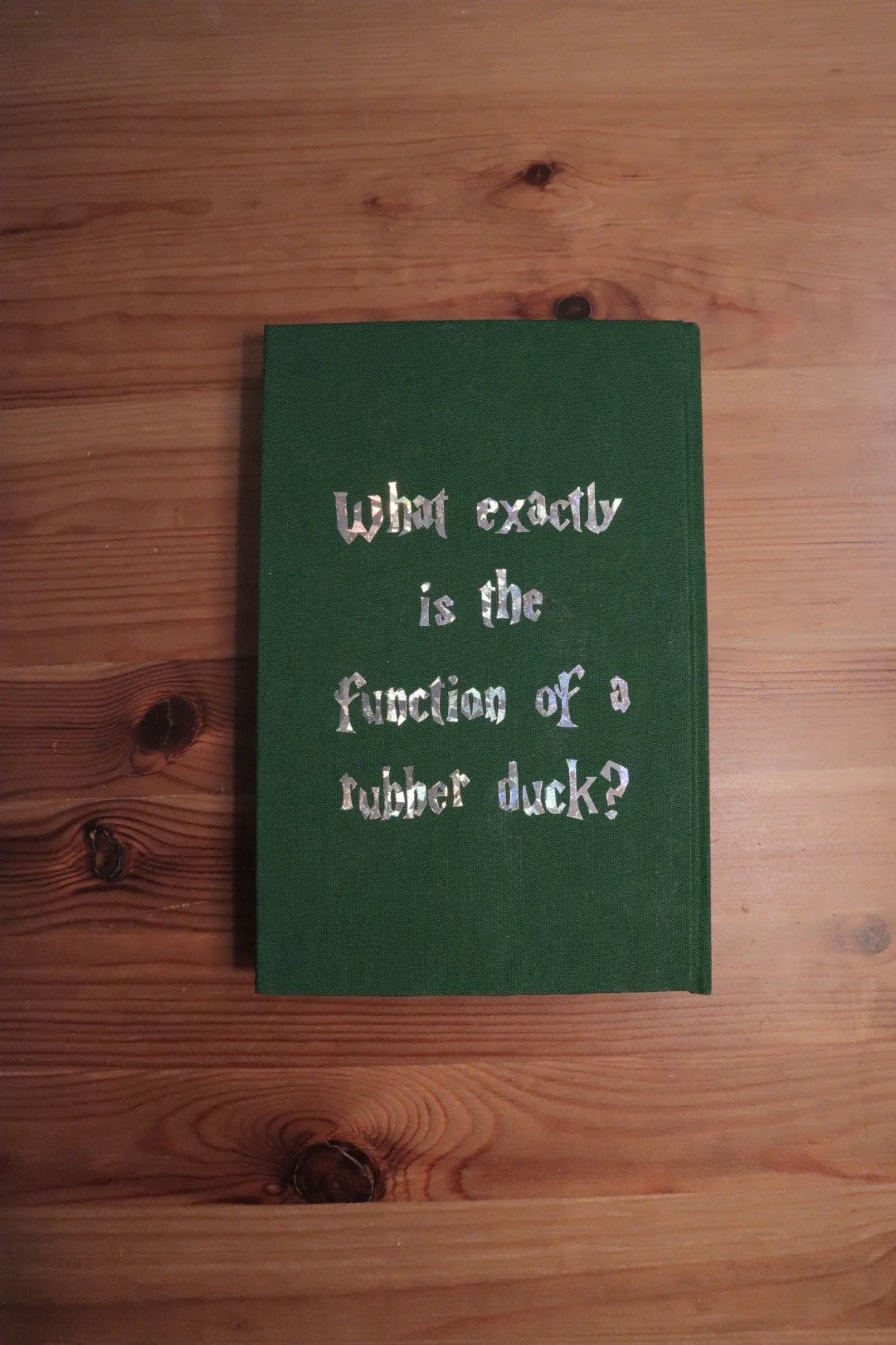 Green book with metallic silver text on a wooden surface. The text reads, 'What exactly is the function of a rubber duck?'.