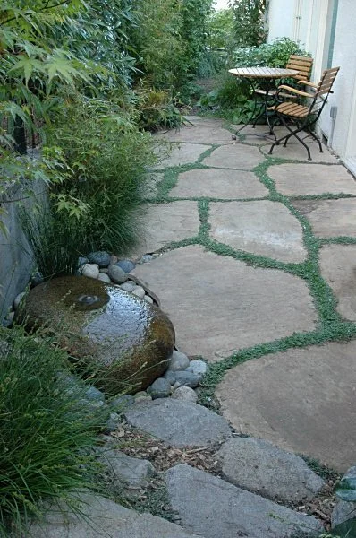 A round stone water fountain next to a tan natural flagstone patio in a small residential back garden with california native plants.