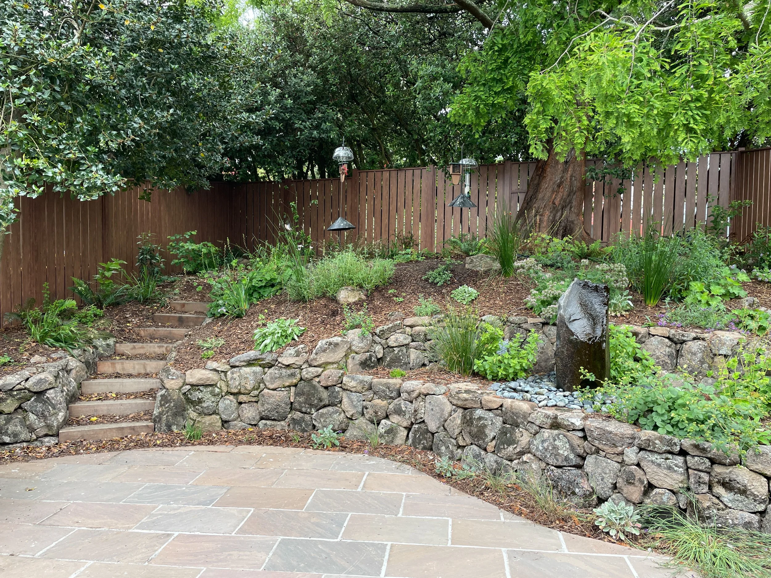 Moss rock walls surround a columnar stone fountain and california native plants behind a stone patio with timber tie steps in this berkeley hills bird garden