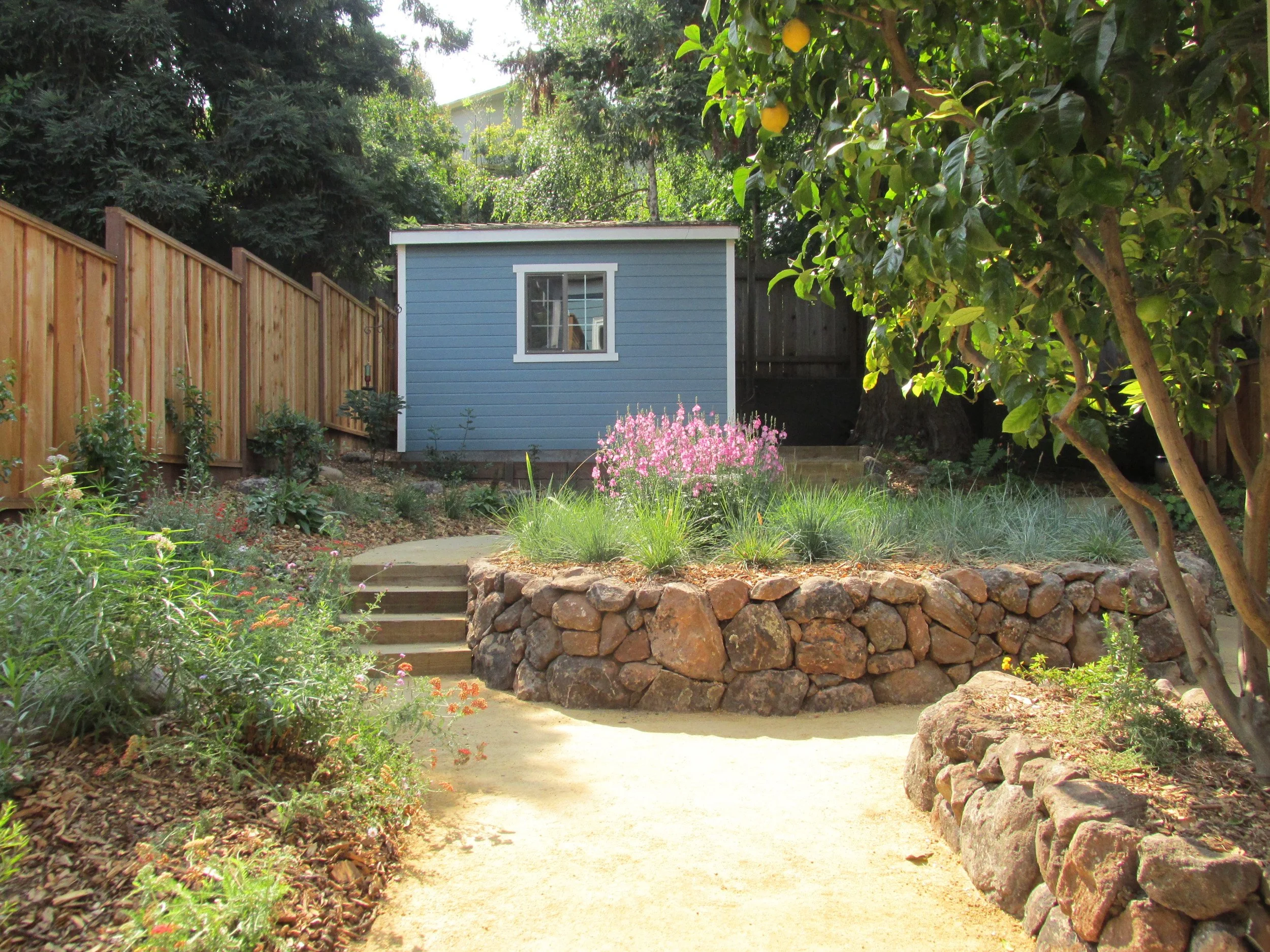 Dry stacked moss rock stone walls curved around california native plant garden bed with tan decomposed granite pathway and timber tie steps.