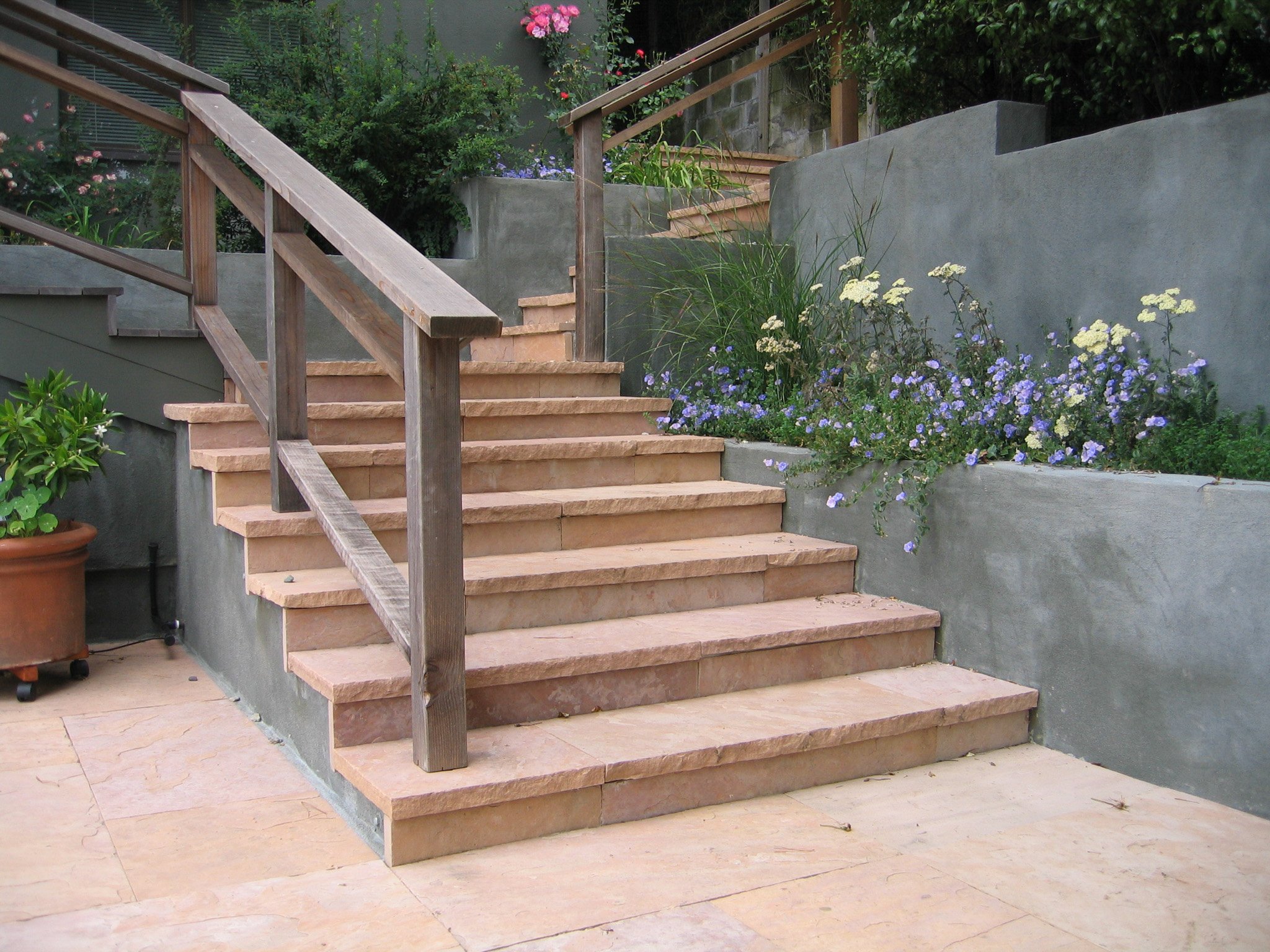 Wood railing down pink stone landscape steps next to grey concrete walls with california native plants yellow yarrow purple 