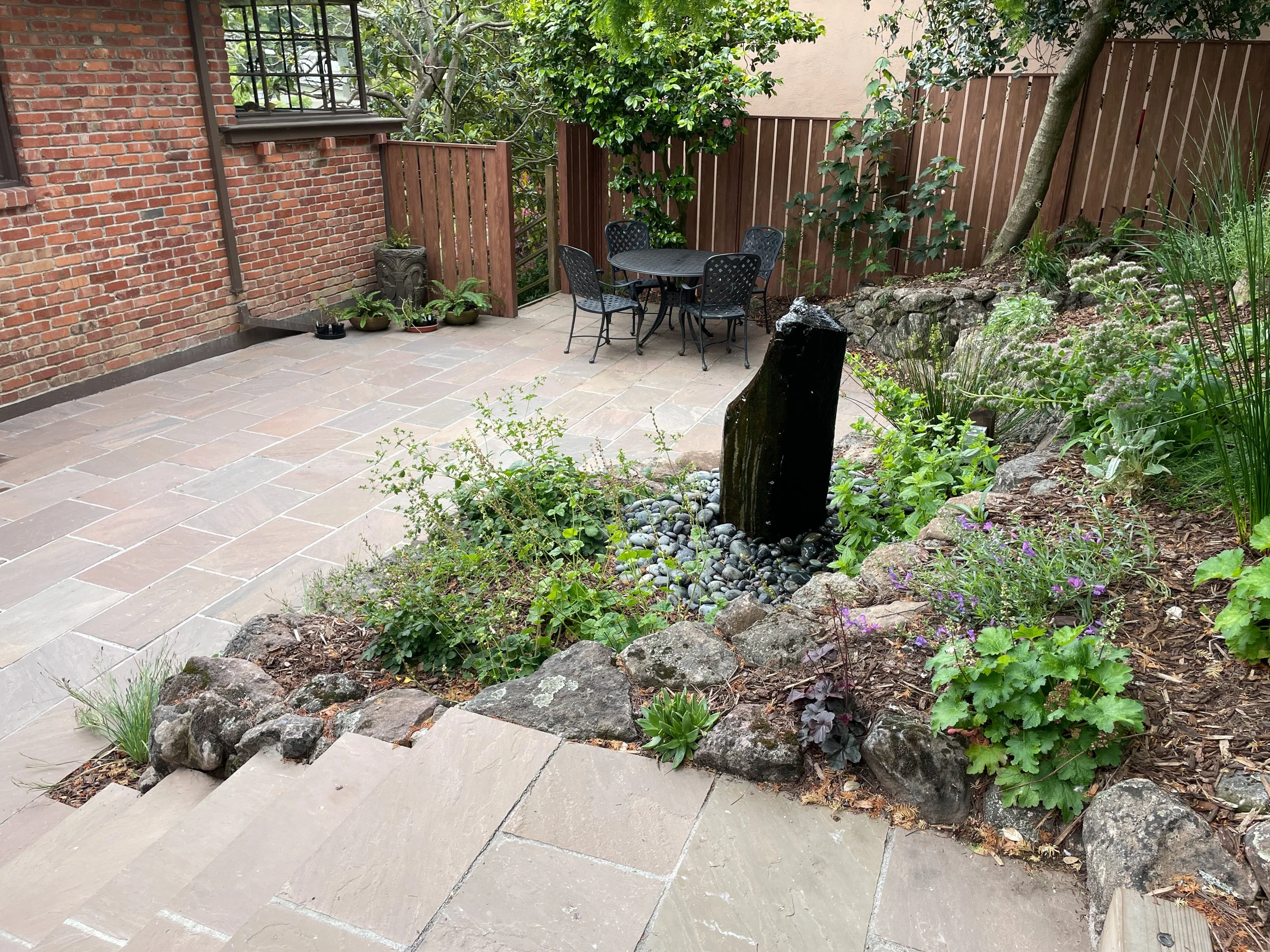 Stone steps and moss rock wall surround a columnar stone fountain with california native plants in this berkeley bird garden