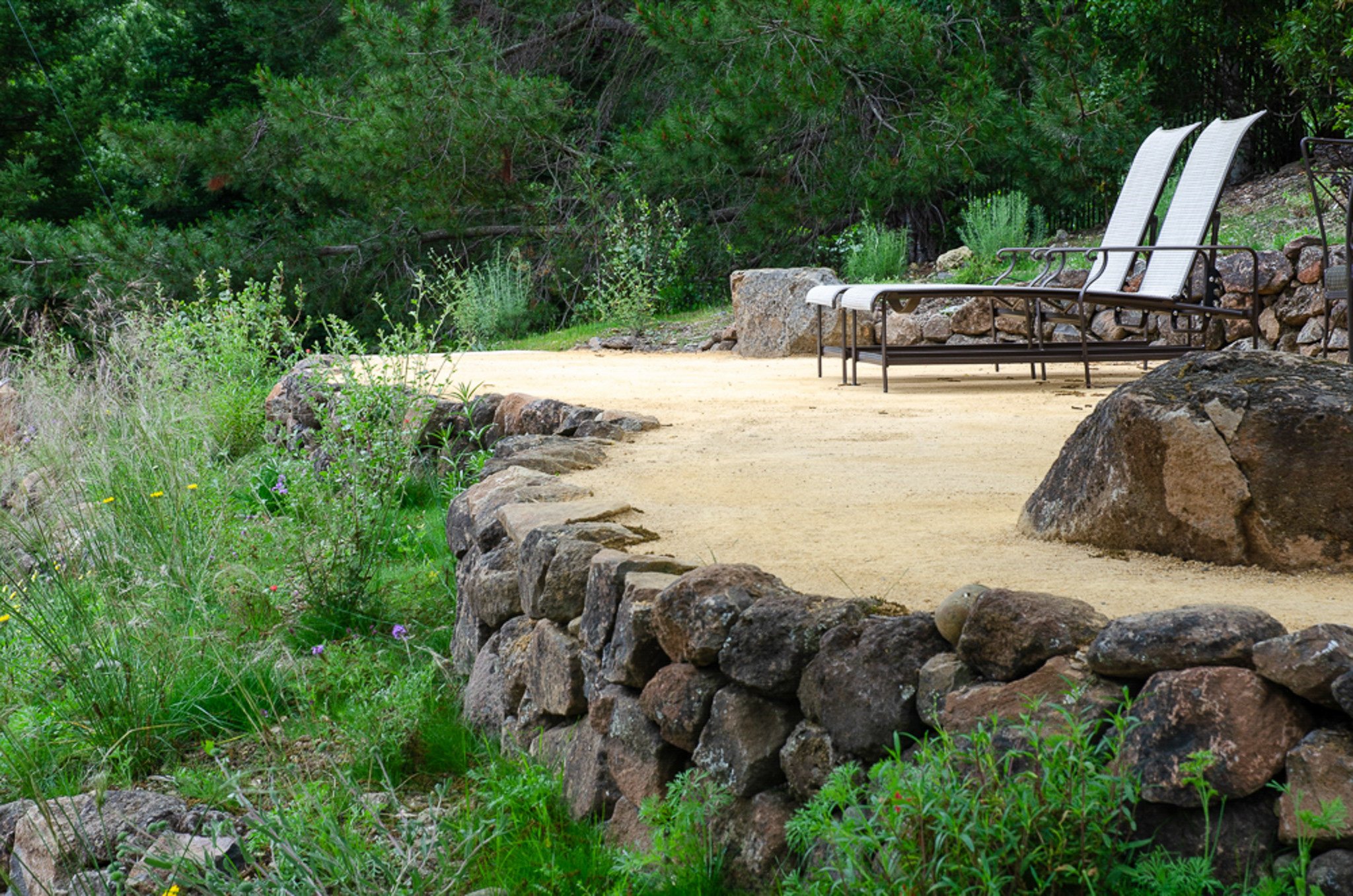 A moss rock dry stacked wood wall with a tan decomposed granite patio with lounge chairs and boulders in a california native plant garden residential landscape.