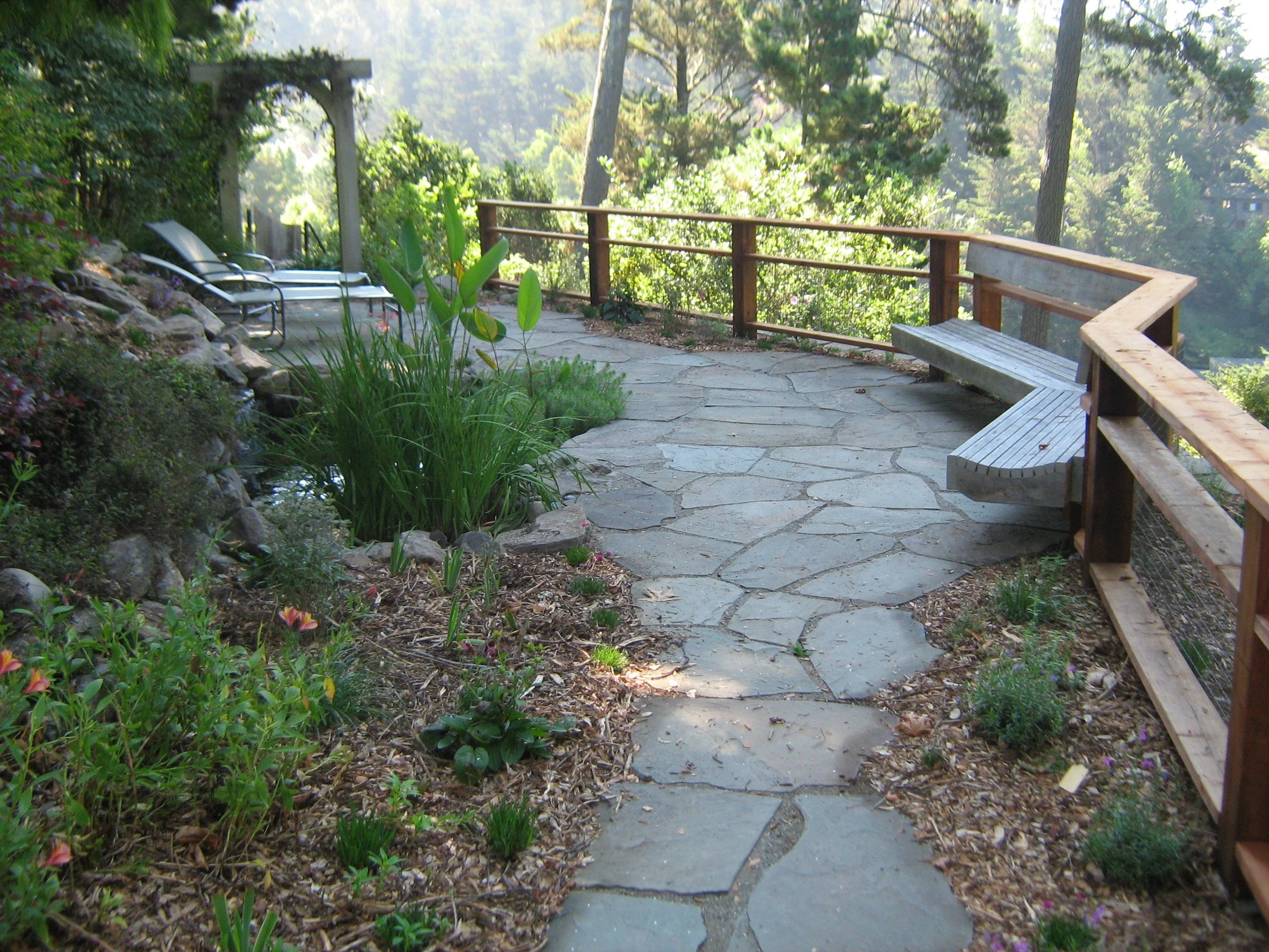 A flagstone patio with a wood railing and built-in wood bench for enjoying the view of the hillside garden and the water feature.