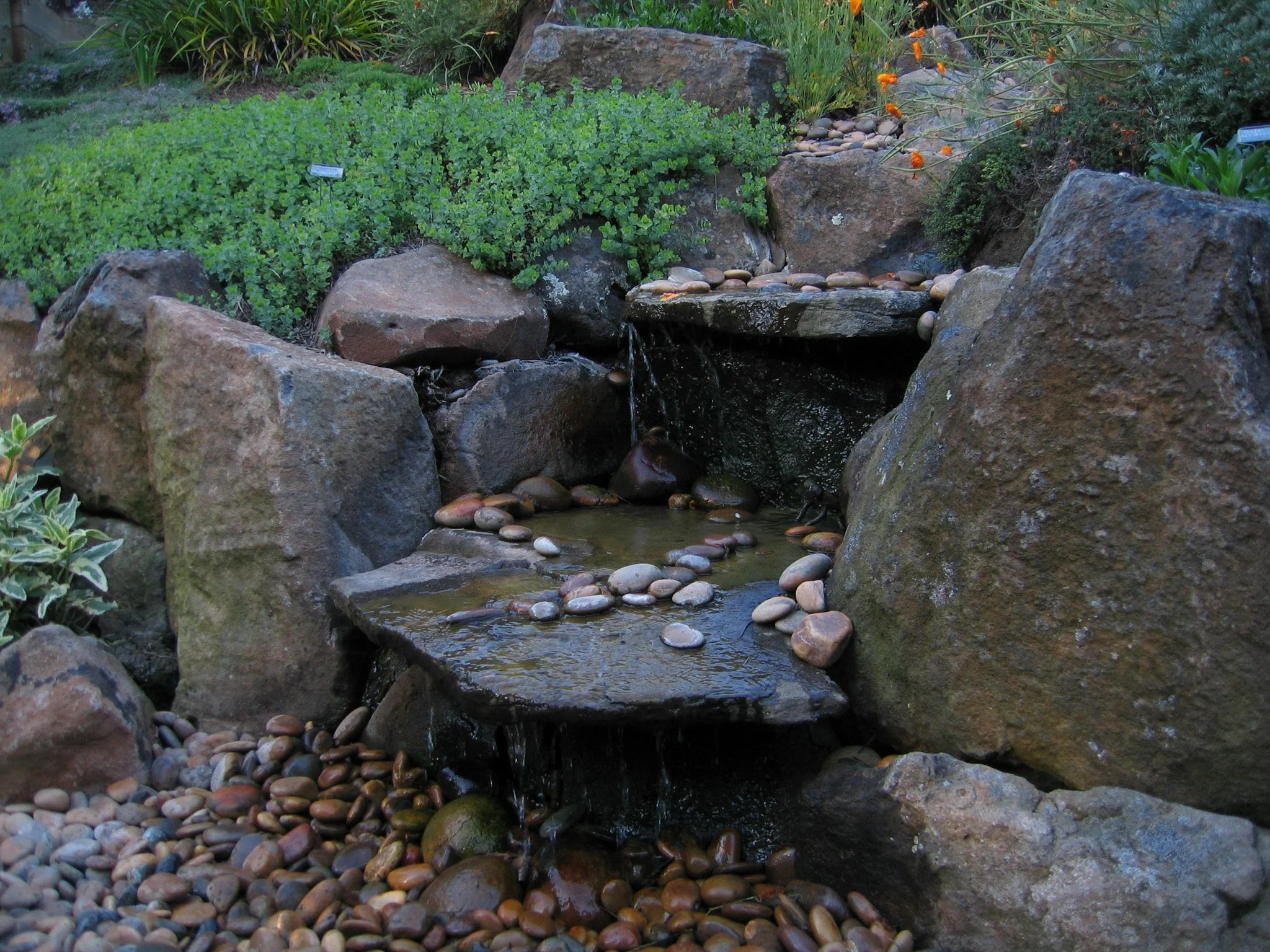Tiered stone waterfall fountain water feature pondless waterfall with gravel base surrounded by california native plants.