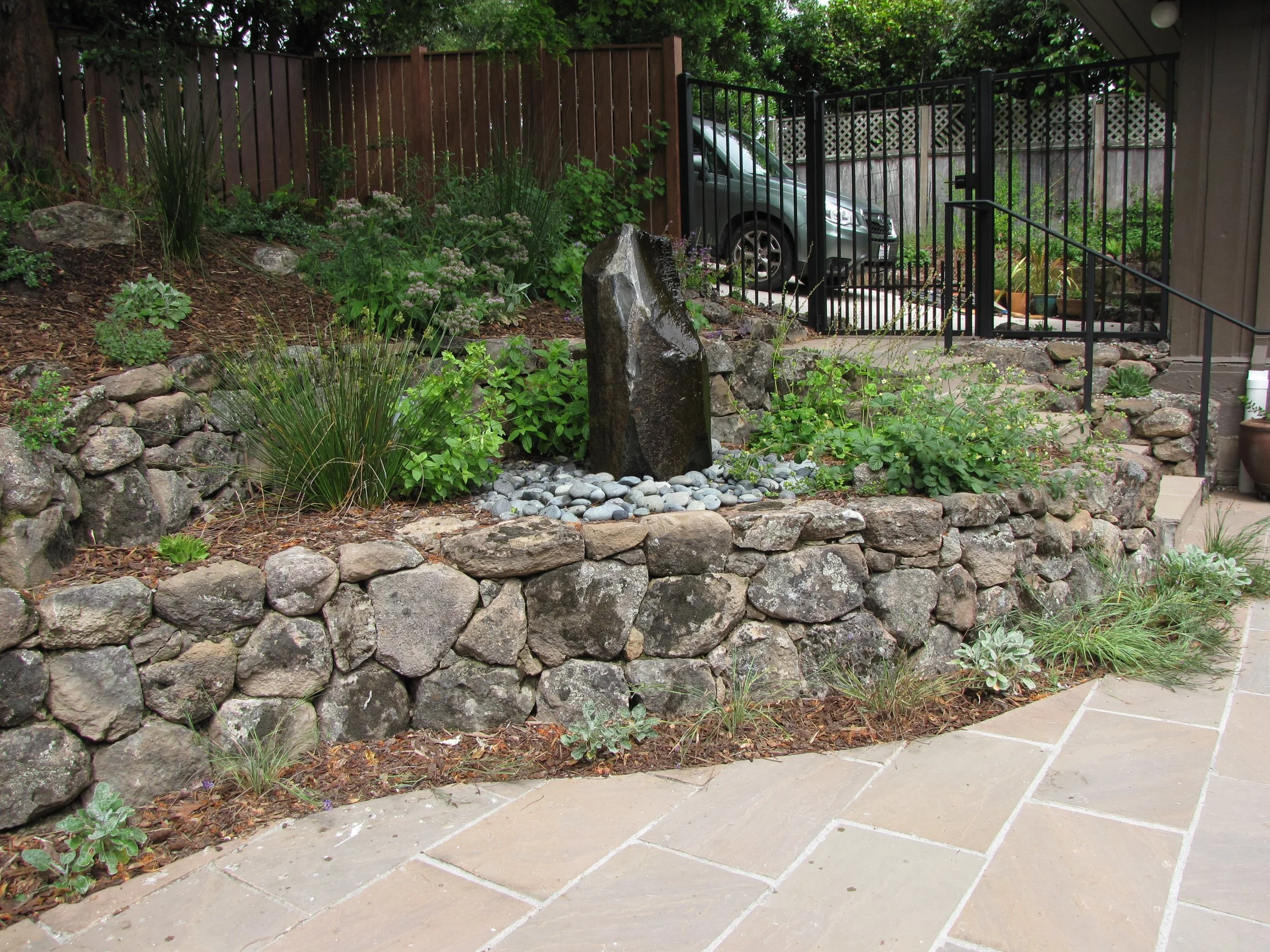 Moss rock walls surround a columnar stone fountain and california native plants behind a stone patio with timber tie steps in this berkeley hills bird garden