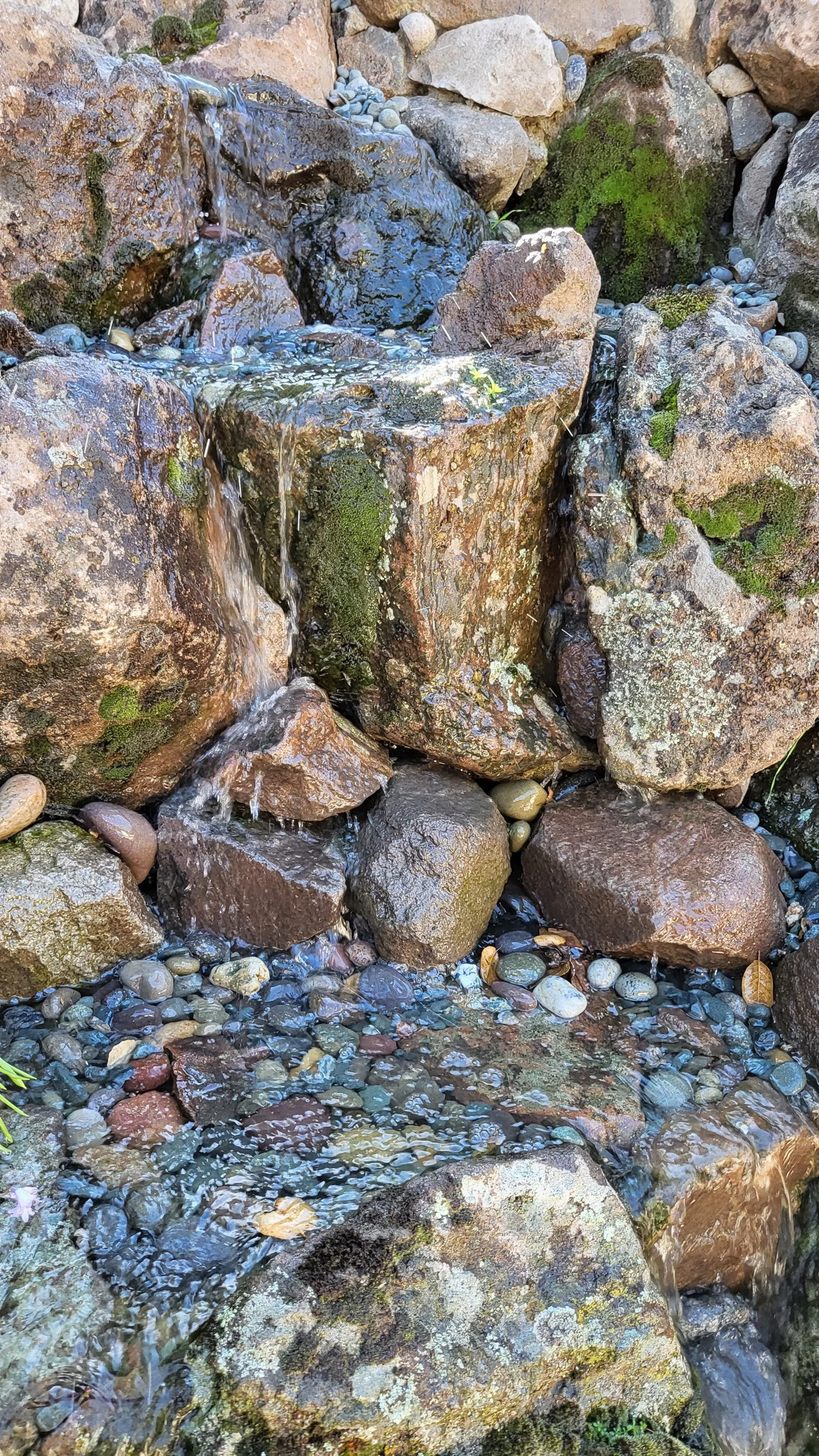 Closeup of stone waterfall natural water feature in residential garden with moss rock and small pebbles.