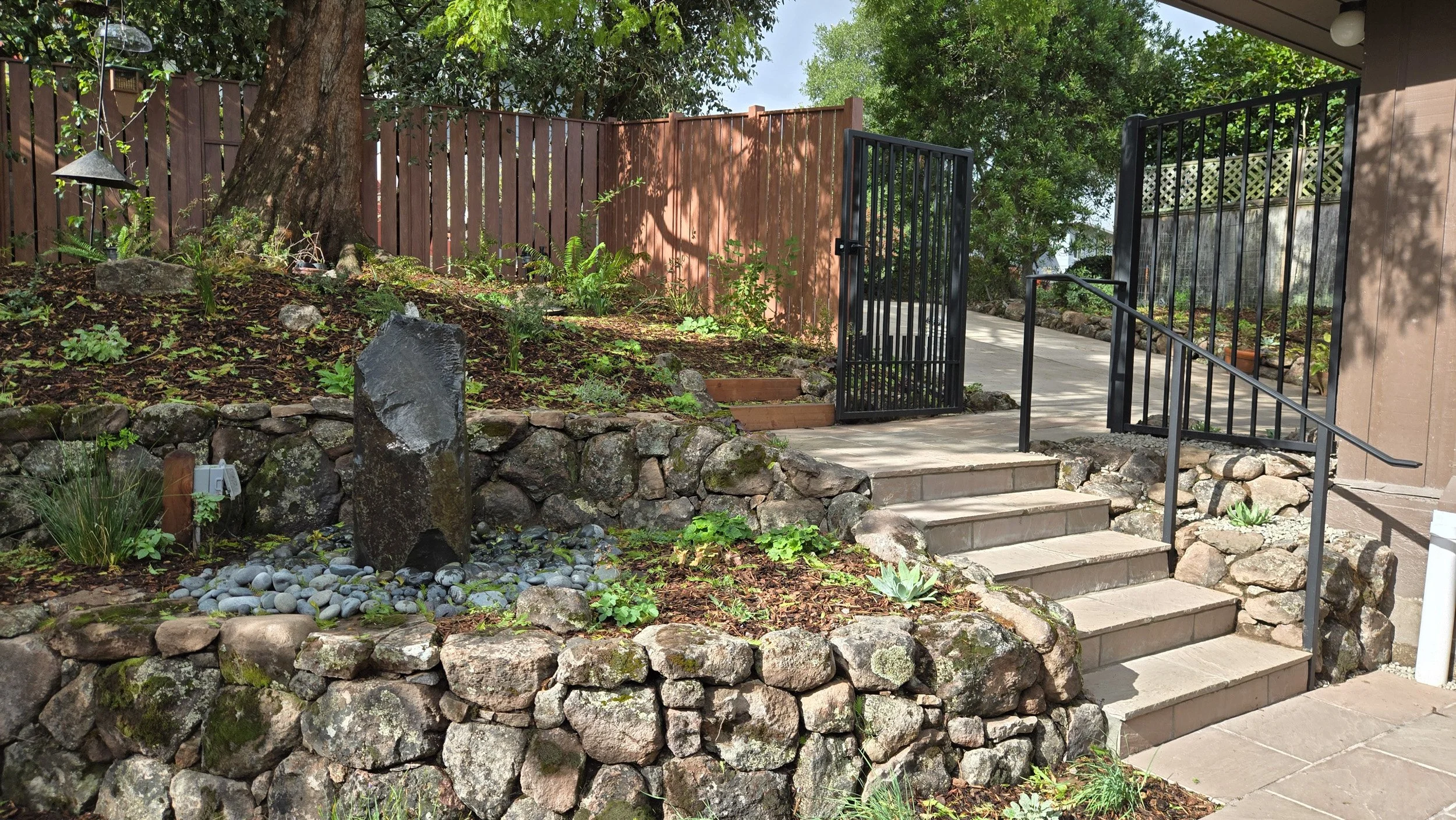 A metal fence and gate entrance protects a california residential home from fire with a rock wall and low california native plantings along stone steps within zone 0.