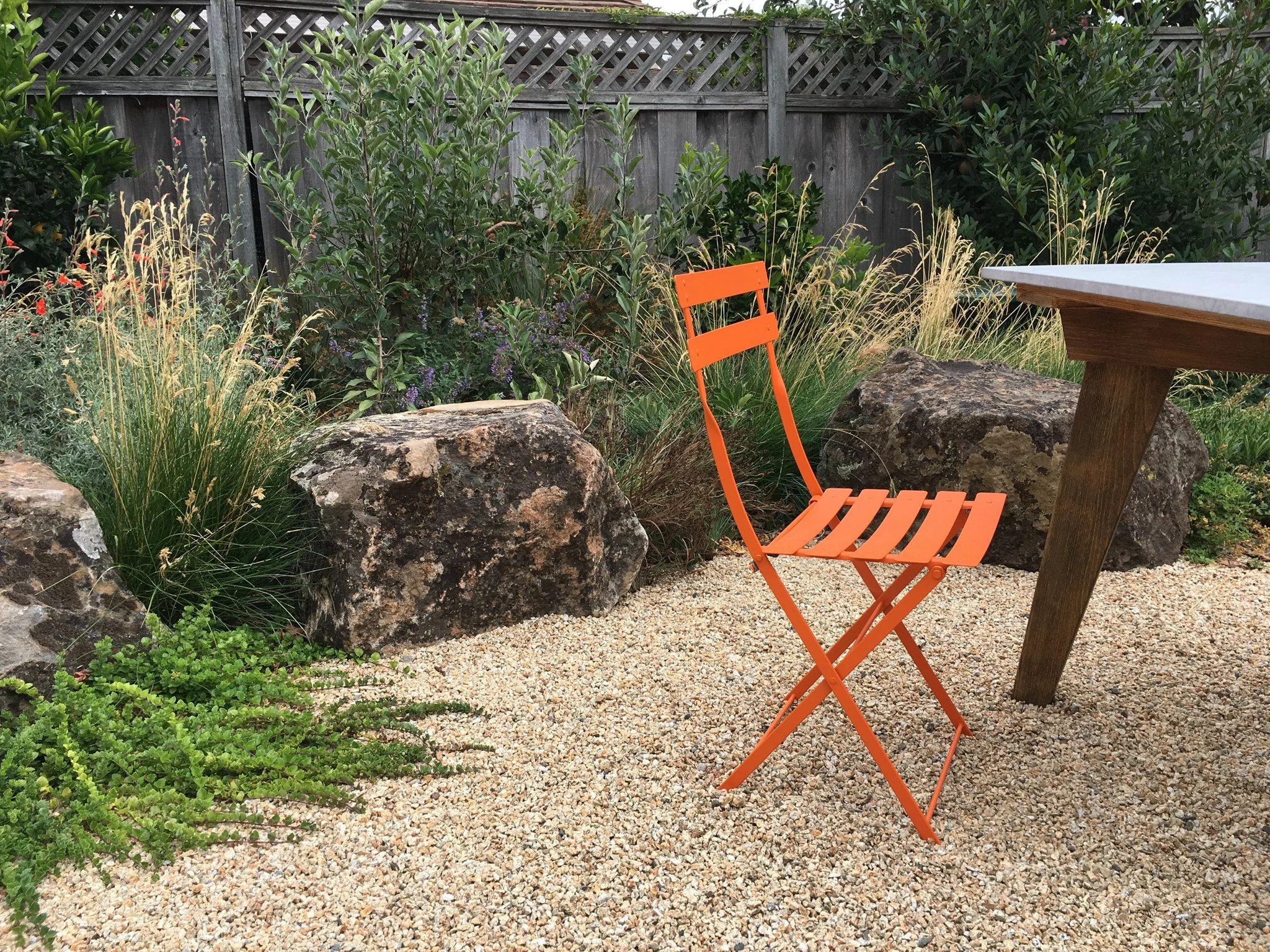 Moss rock boulder next to gravel patio with orange garden chair and native perennial plantings