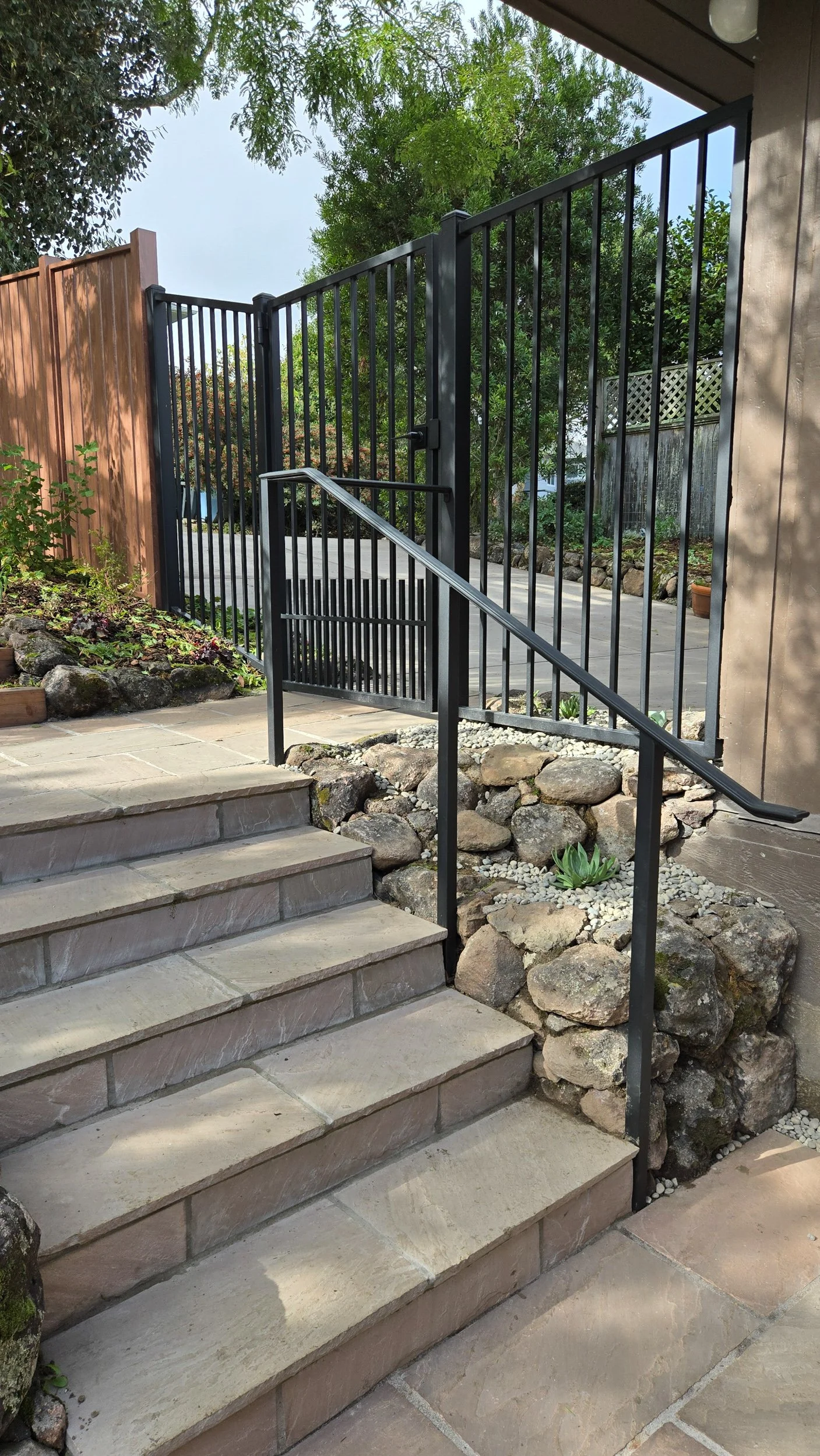 A stone patio and steps with metal fencing and a metal railing in a residential garden with california native plants with stone rock walls