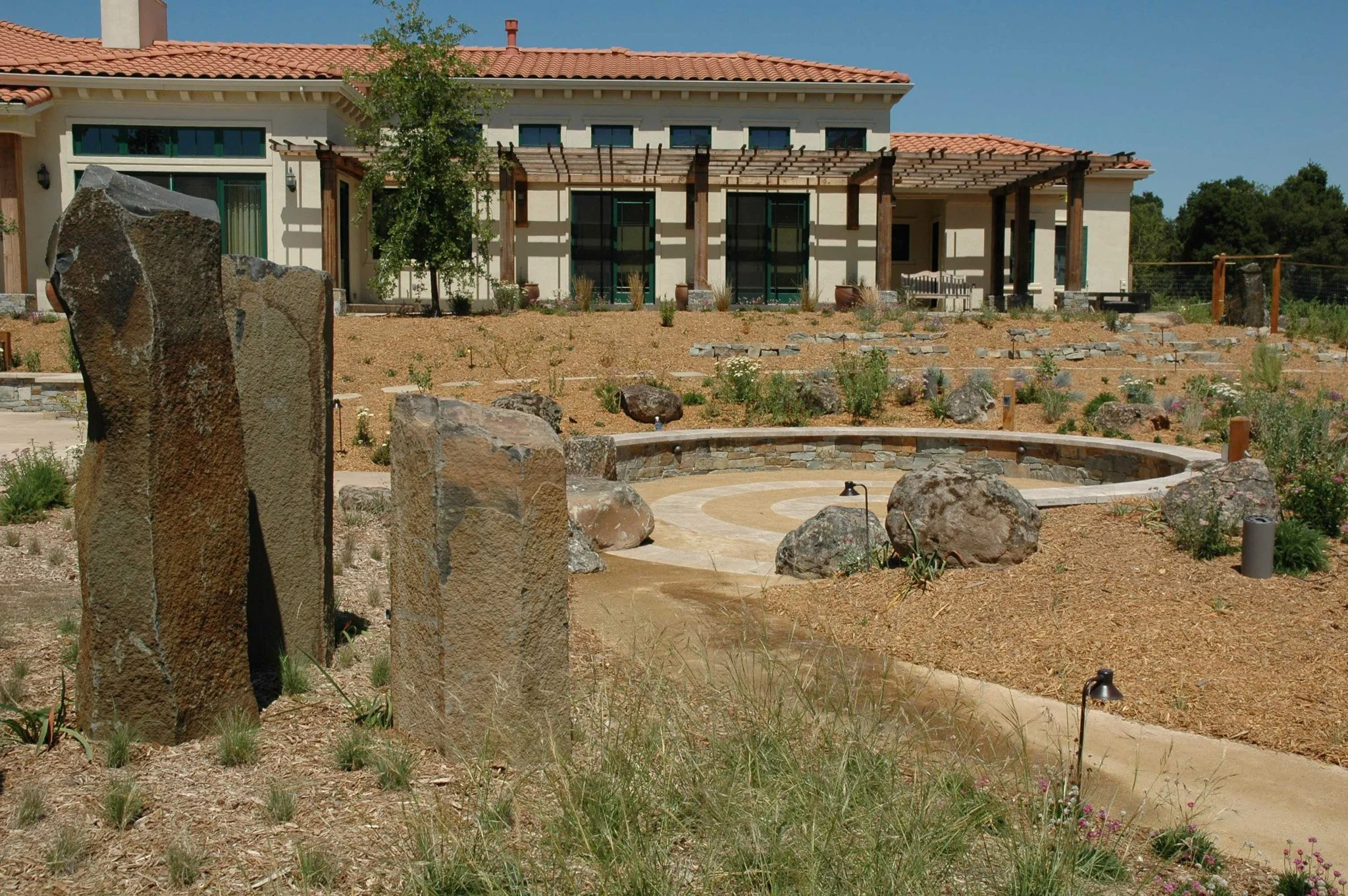 Columns of rock in a trio near a circular stone patio with california native plant grasses mulch and a pergola.