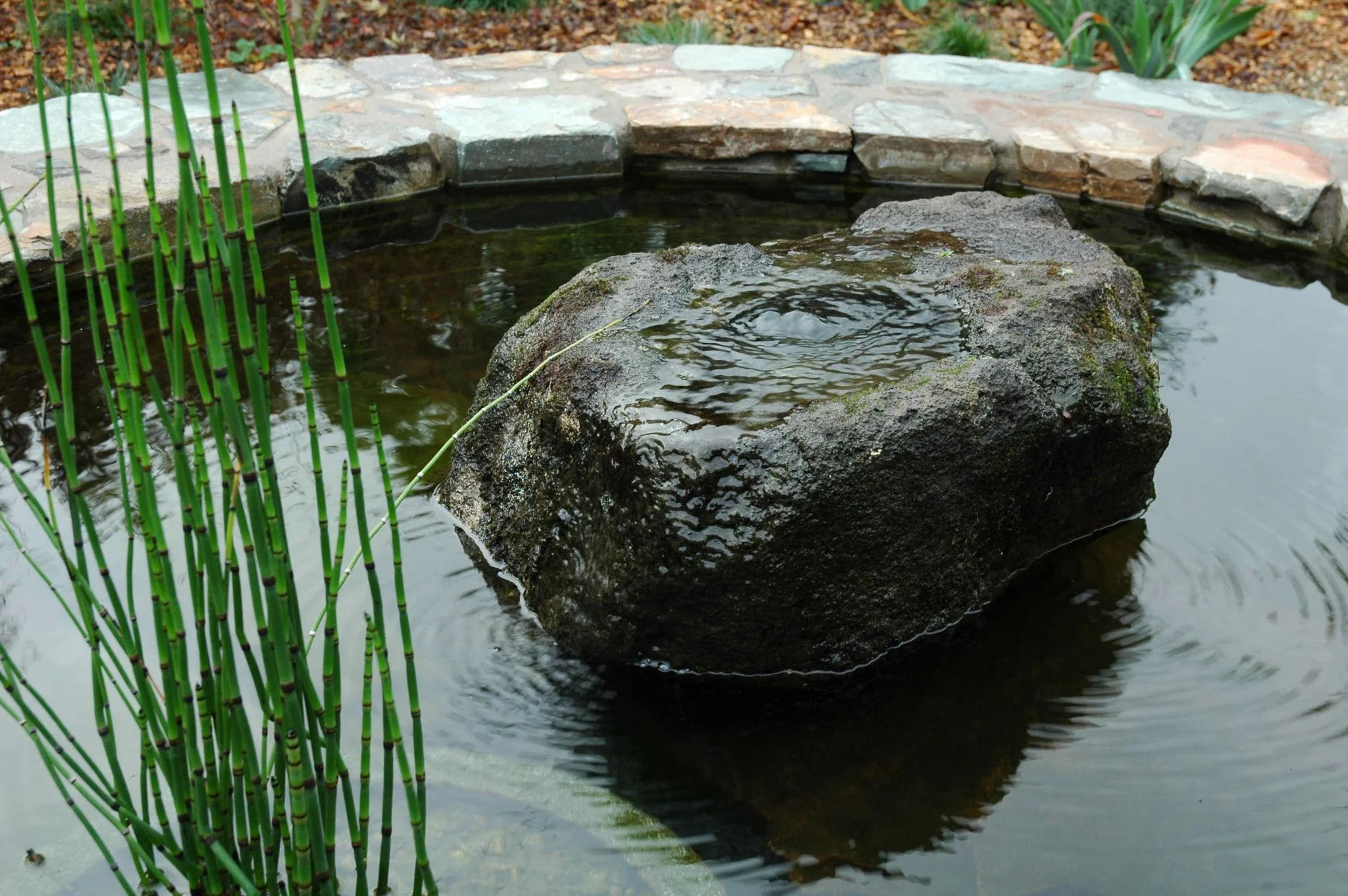 A large boulder fountain in a circular pond surrounded by a rock wall.
