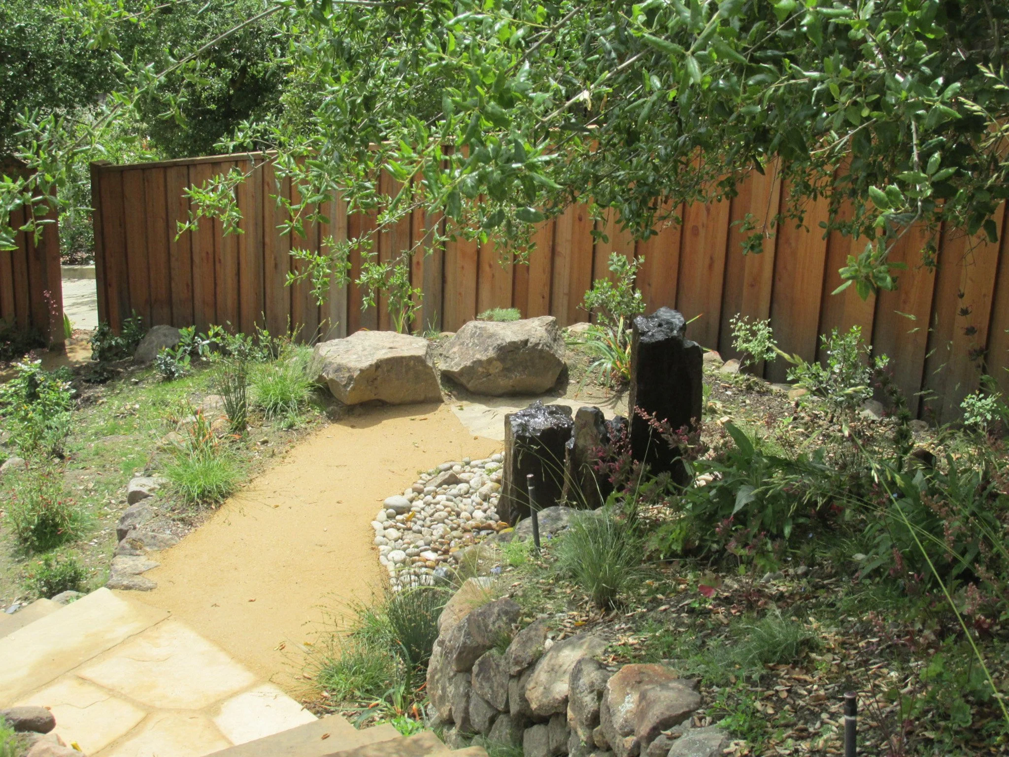 A trio of stone columns form a natural rock water feature fountain with large boulder seats and a decomposed granite patio near flagstone in a residential backyard.