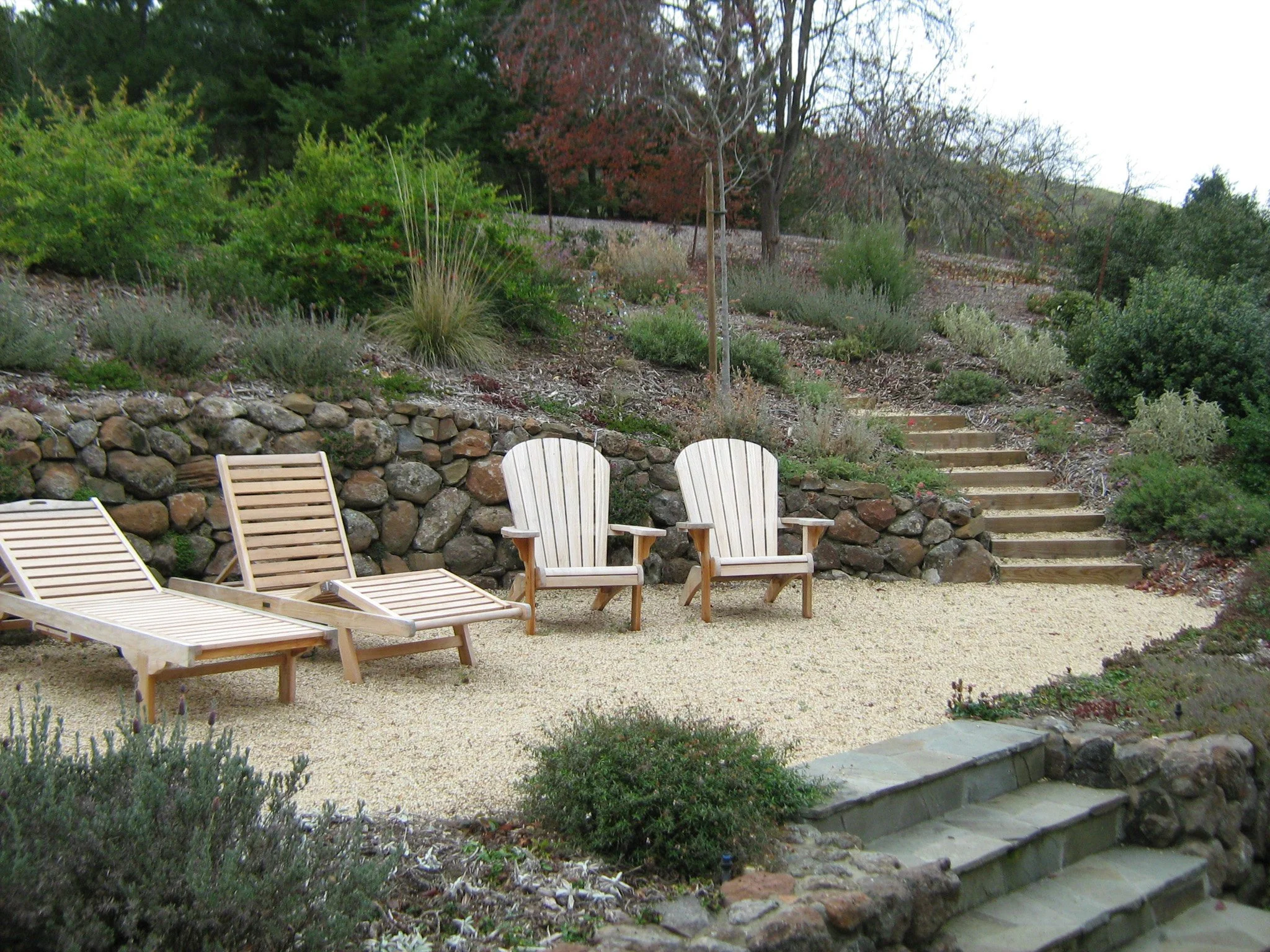  Natural wood lounge and adirondack chair seating on a tan gravel patio with stone steps and timber tie steps with a moss rock dry stacked stone wall in a california native plant garden.