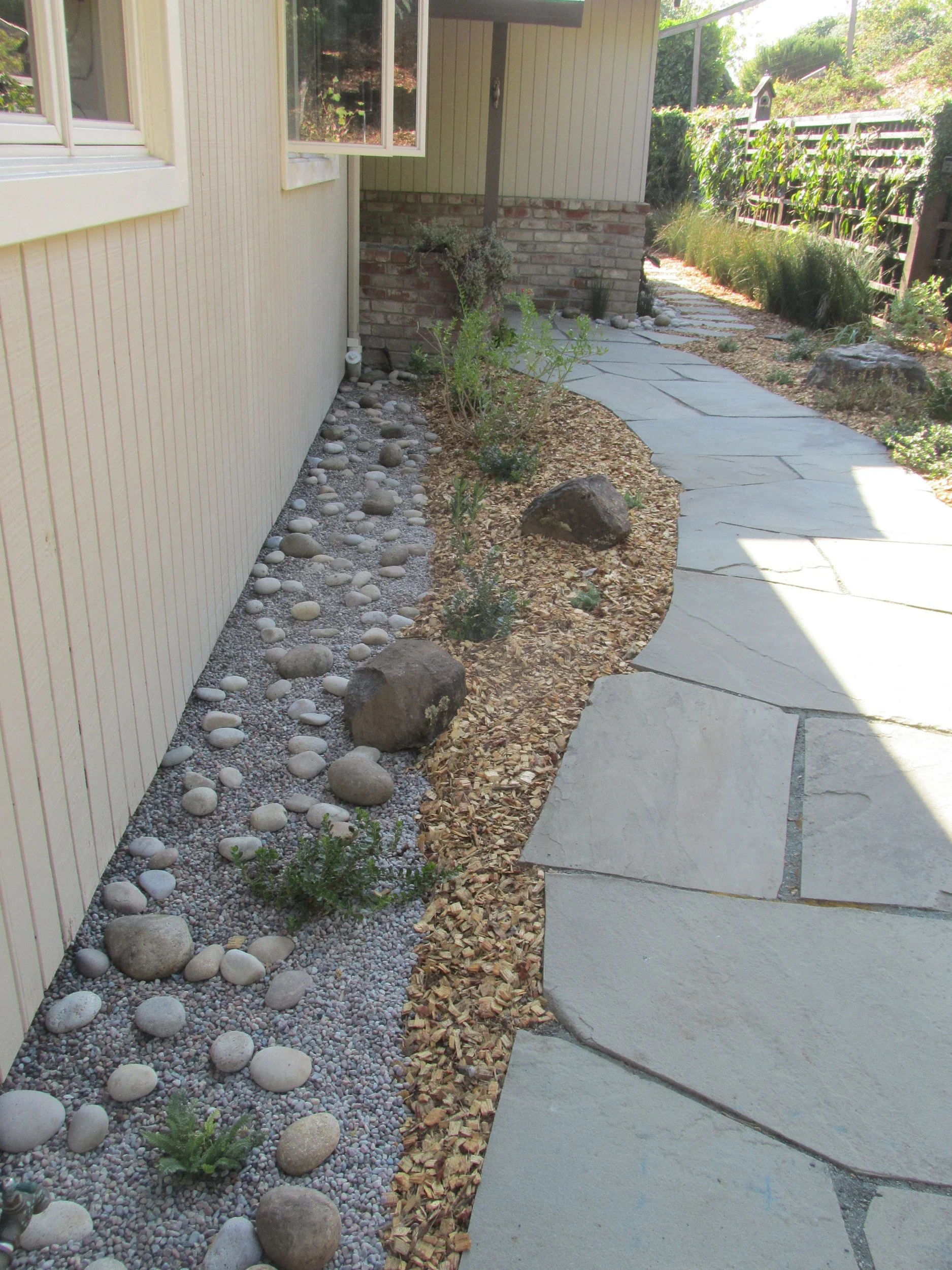 A rock and gravel barrier along a residential home creates a firescaping buffer next to a curving grey flagstone pathway and low plantings