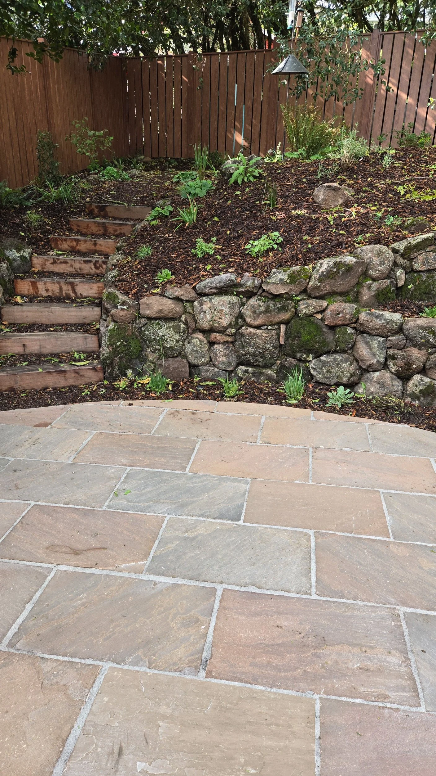 Tan geometric paver patio next to timber tie steps and a dry stacked moss rock wood wall in a california native plant garden.