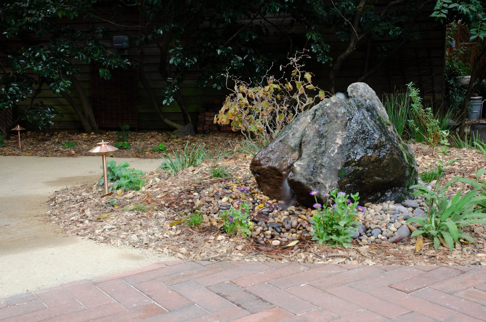 A triangular stone boulder rock fountain in a california native garden with herringbone brick paving and modern copper path lighting.