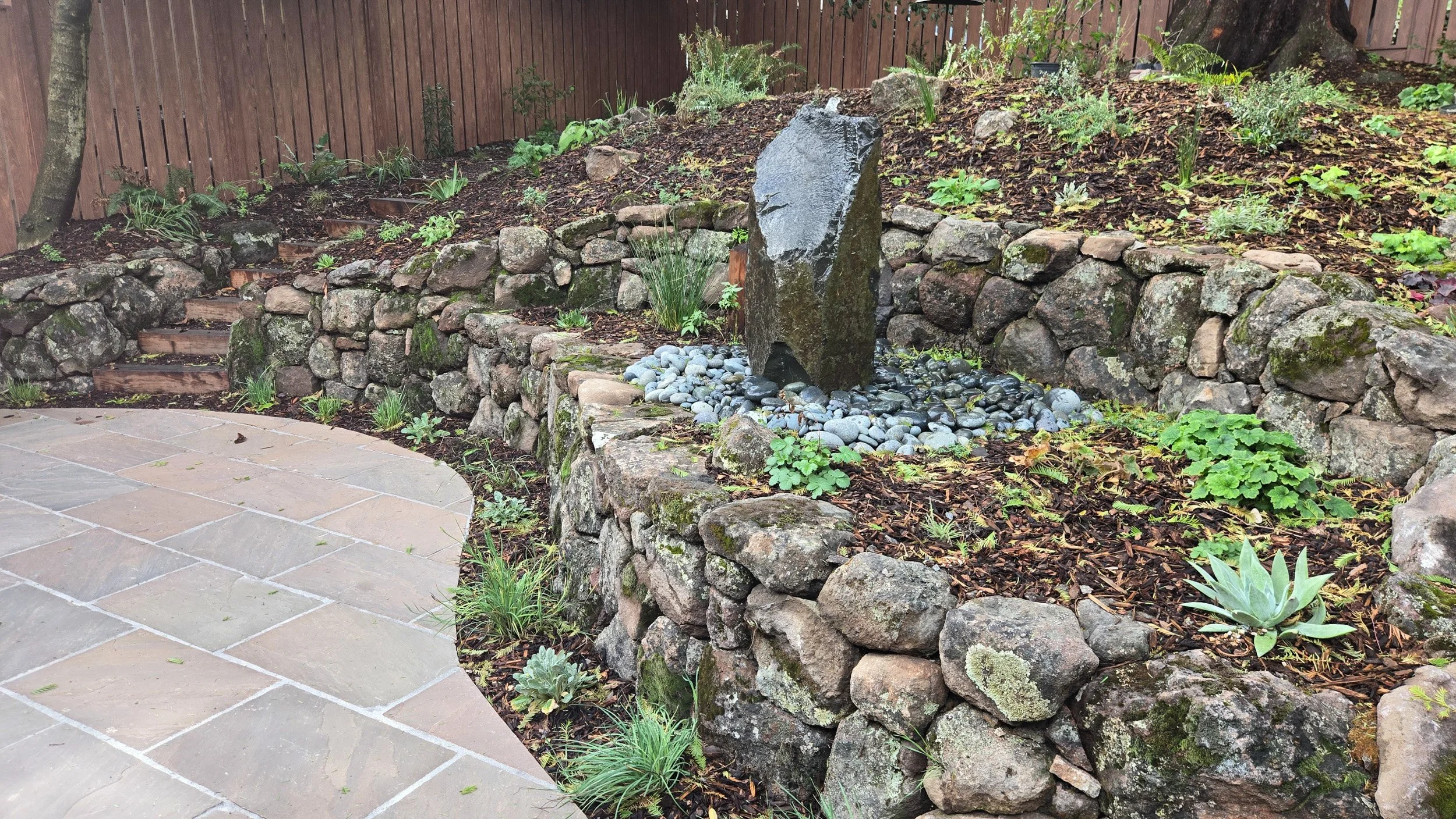 A tall vertical rock fountain on a terraced moss rock wall residential garden with california native plants and a formal stone paver patio.