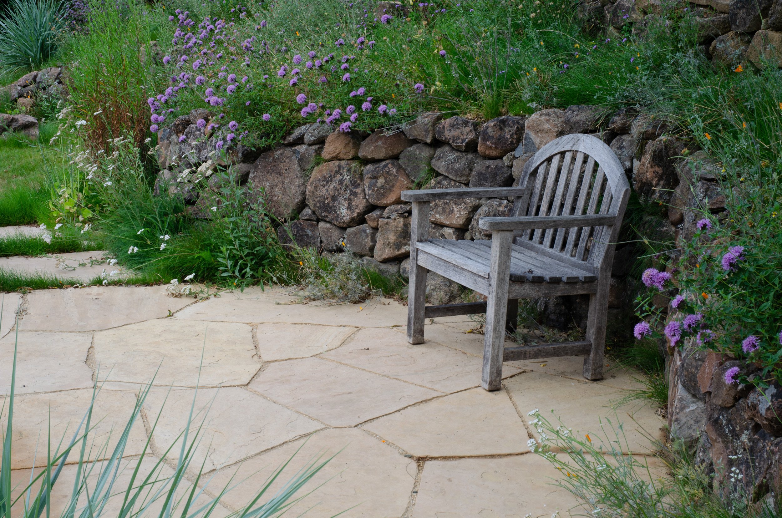 A wood bench tucked into an alcove of the natural stone wall on a flagstone patio, looking out over this hillside wildlife garden.