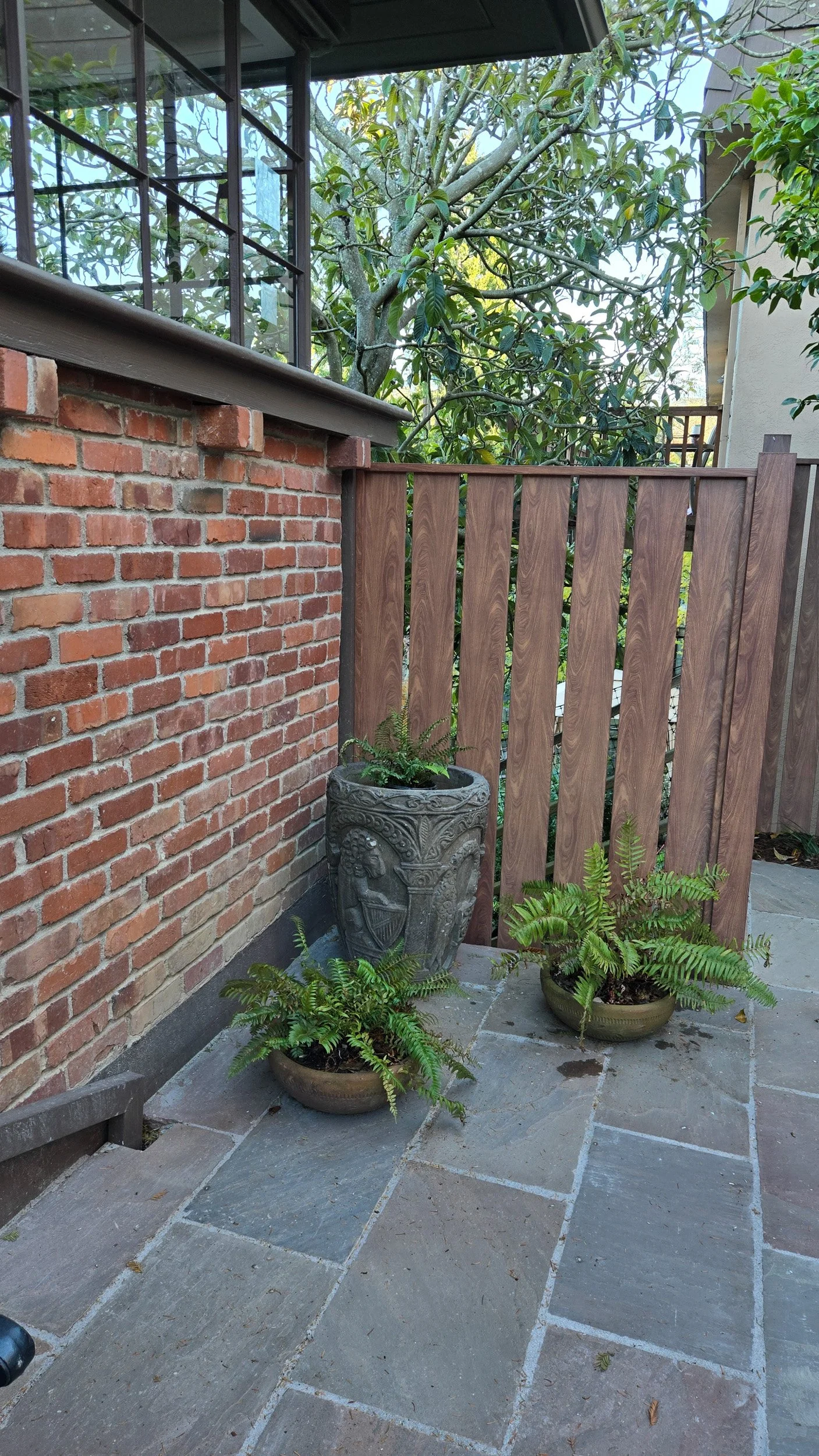 A wood-look metal fence panel next to a brick wall on a stone patio with potted ferns