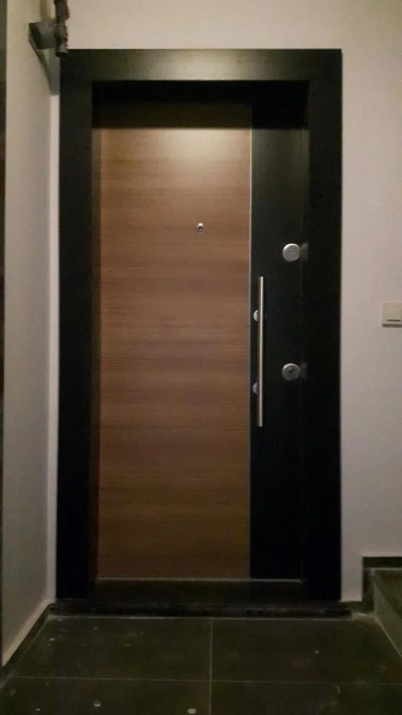 A front door with a wood finish, framed by black trim, featuring a peephole, a handle, and security locks, with a white wall on the right and a dark floor.