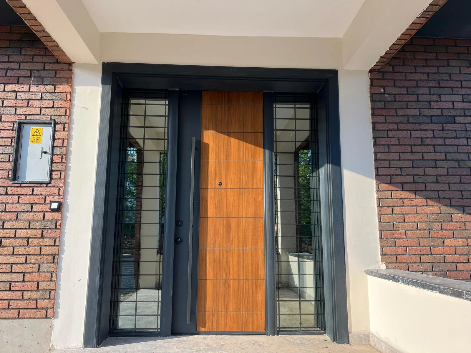 Modern front door with wood paneling, black metal frame, glass sides, brick and white wall facade.