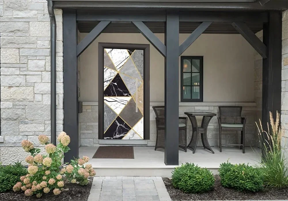 Ön verandada kapıda siyah ve altın rengi geometrik desenler, koyu renk hasır masa ve sandalyeler, taş duvar, açık pembe çiçeklerle dolu küçük bir bahçe ve gri kaldırım taşları bulunmaktadır.