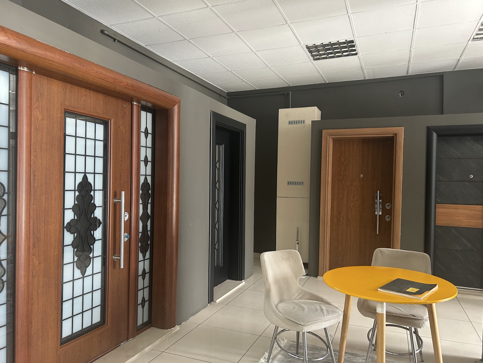 İç mekânda, siyah işlemeli cam panellerle süslenmiş iki ahşap kapı, gri ve siyah duvar detayları, bej bir sandalye ve üzerinde siyah bir kitap bulunan sarı yuvarlak bir masa yer alıyor.