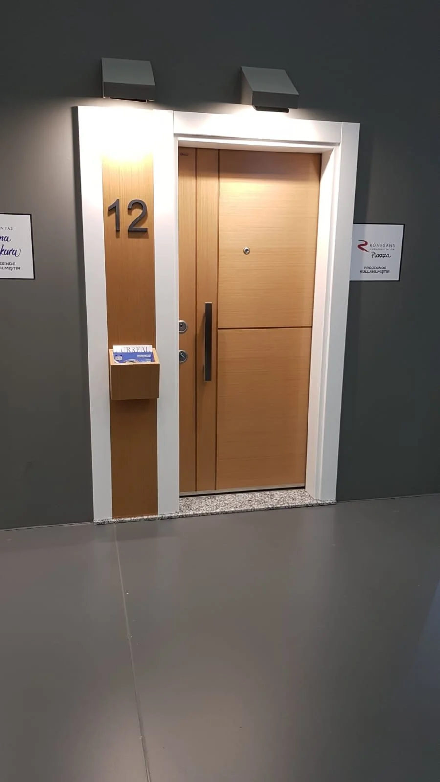 Wooden elevator door with the number 12 on a gray wall, a small mailbox attached, and signs on either side of the door.