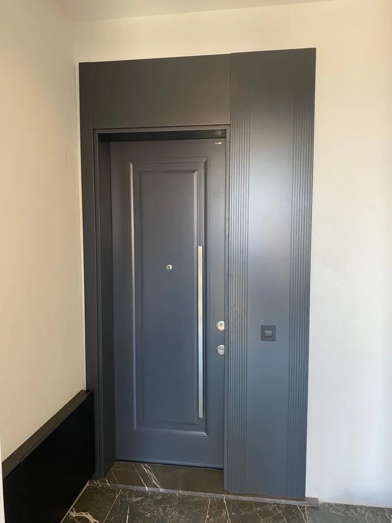 A modern dark gray front door with vertical panel design and a long metal handle, installed in a wall with matching vertical panel accents, next to a light-colored wall with a square doorbell button and a black baseboard on the left, with dark marble