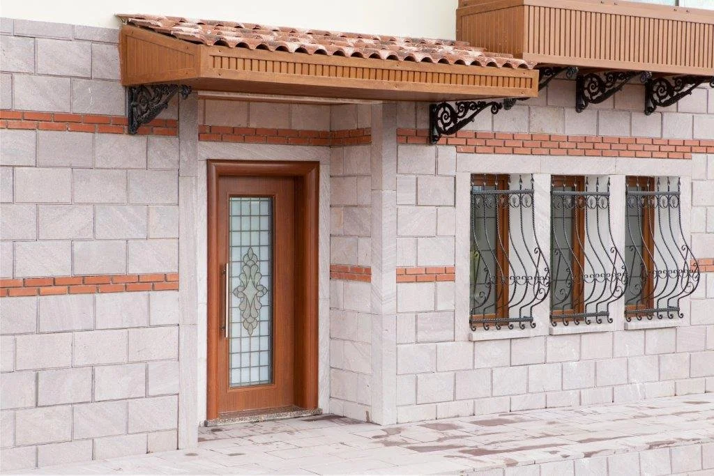 Front entrance of a house with a wooden door with decorative glass, three windows with ornate metal grilles, stone walls with brick accents, and a small tiled porch.