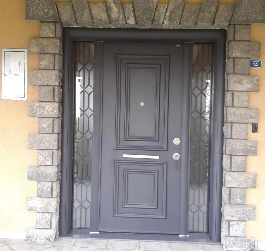 Dekoratif panellerle süslenmiş siyah ön kapı, geometrik desenli metal ızgaralı yan camlar, taş çerçeveyle çevrili, bej dış duvar, mavi levha üzerinde 14 numaralı ev.