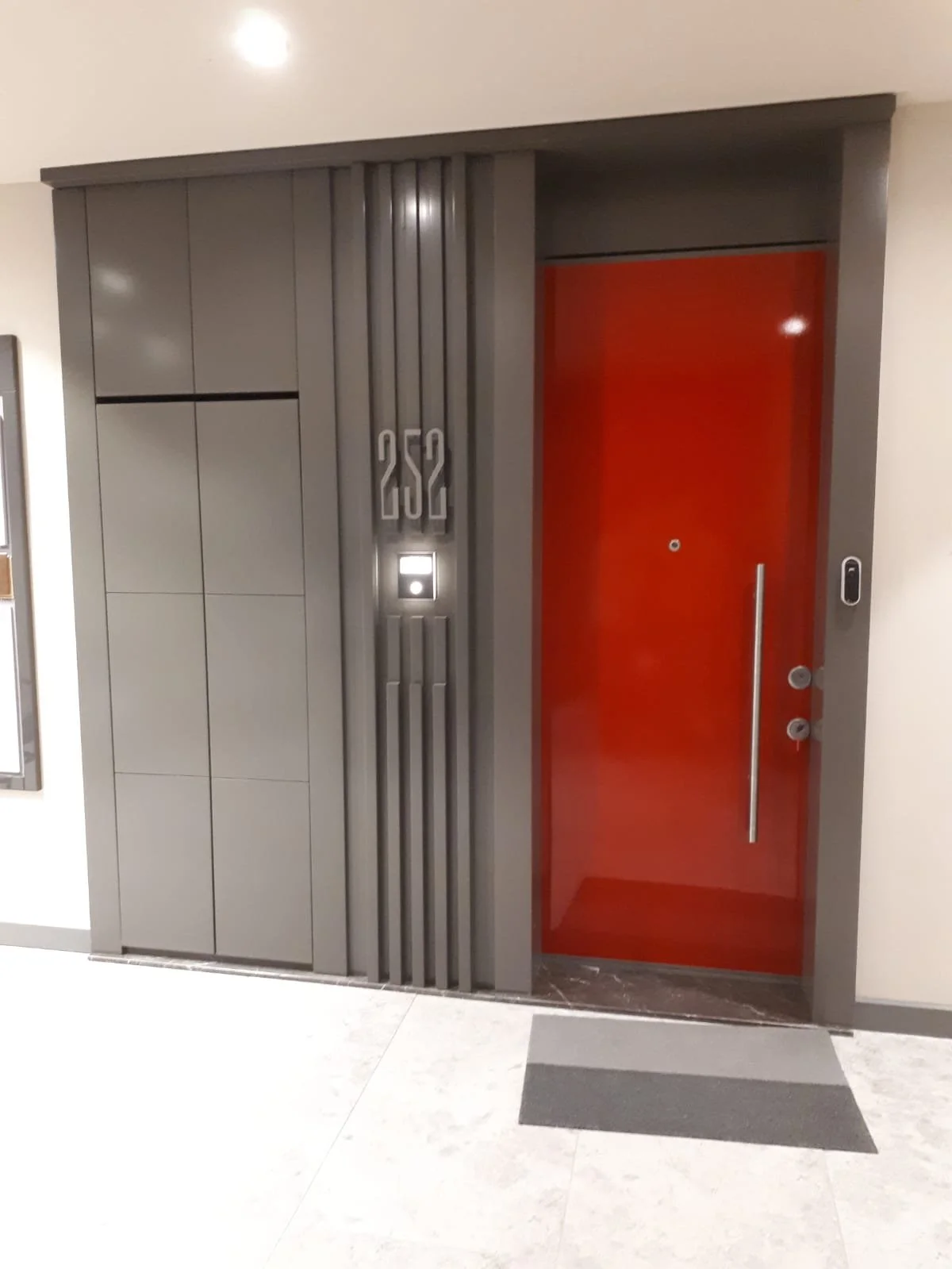 Modern building entrance with grey paneling, grey metallic numbers 252, a door with a bright red interior, and a small access control device on the wall.
