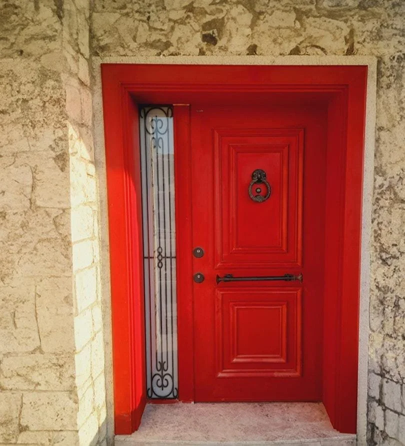 Red front door with decorative black iron handle and a window with black iron grille on the left side, set in a stone wall.