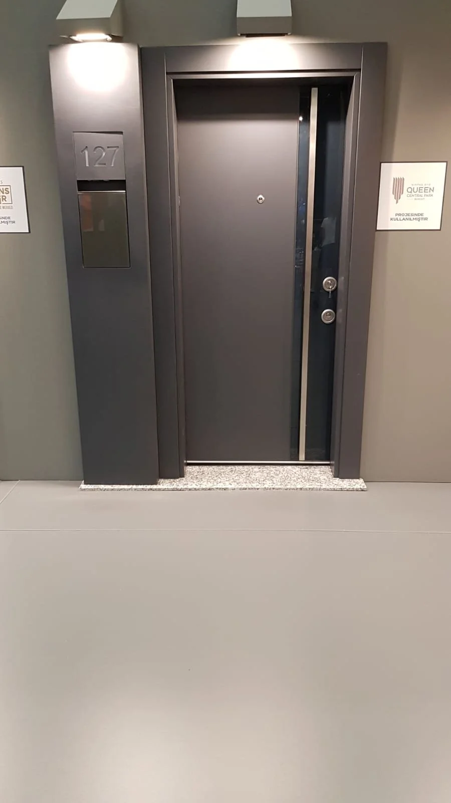 A black security door with a peephole, two locks, and a vertical metal strip. To the left of the door is a gray wall with the number 127 and a small window. To the right, there is a white sign with a logo and some text.