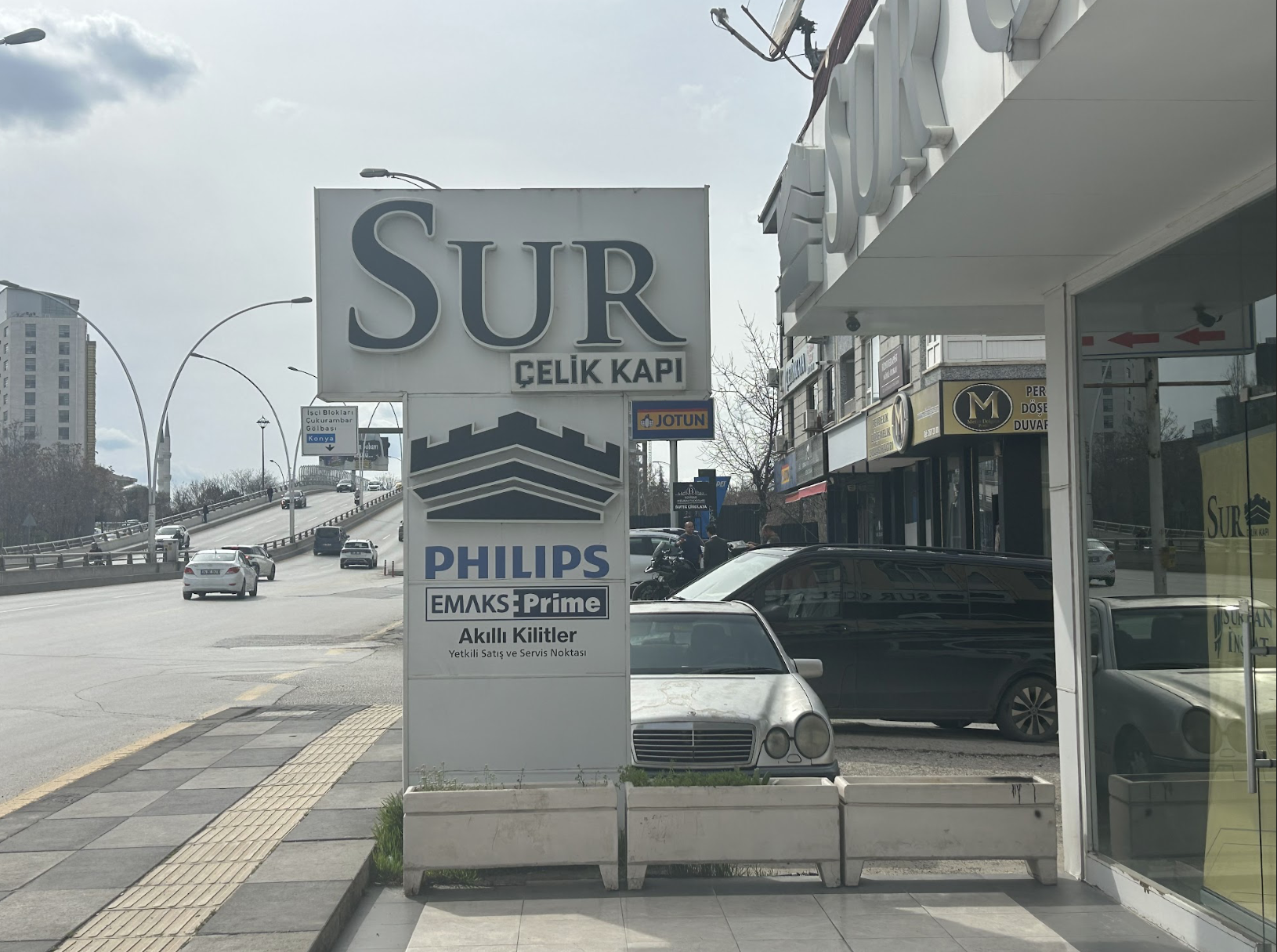 Kaldırım ve park halindeki arabaların yanında, Sur Çelik Kapı, Philips ve EMKS Prime reklamlarının yer aldığı büyük bir tabelanın bulunduğu sokak manzarası. Arka planda binalar, yolda arabalar ve bulutlu bir gökyüzü görülüyor.
