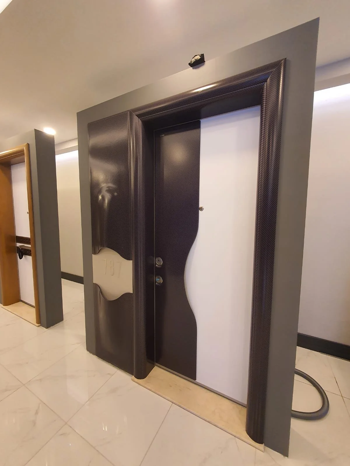 Elevator closet with black carbon fiber design panels and a white door, situated in an indoor hallway with white tiled floor and nearby wooden door.