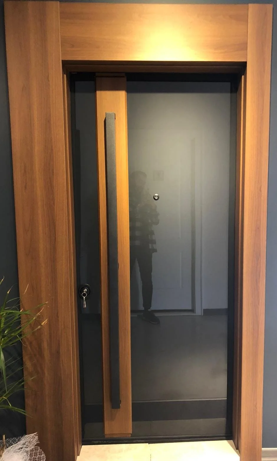 A modern elevator with a glass door framed in wood, a black handle, and a key lock, with a reflection of a person taking the photo visible on the glass.