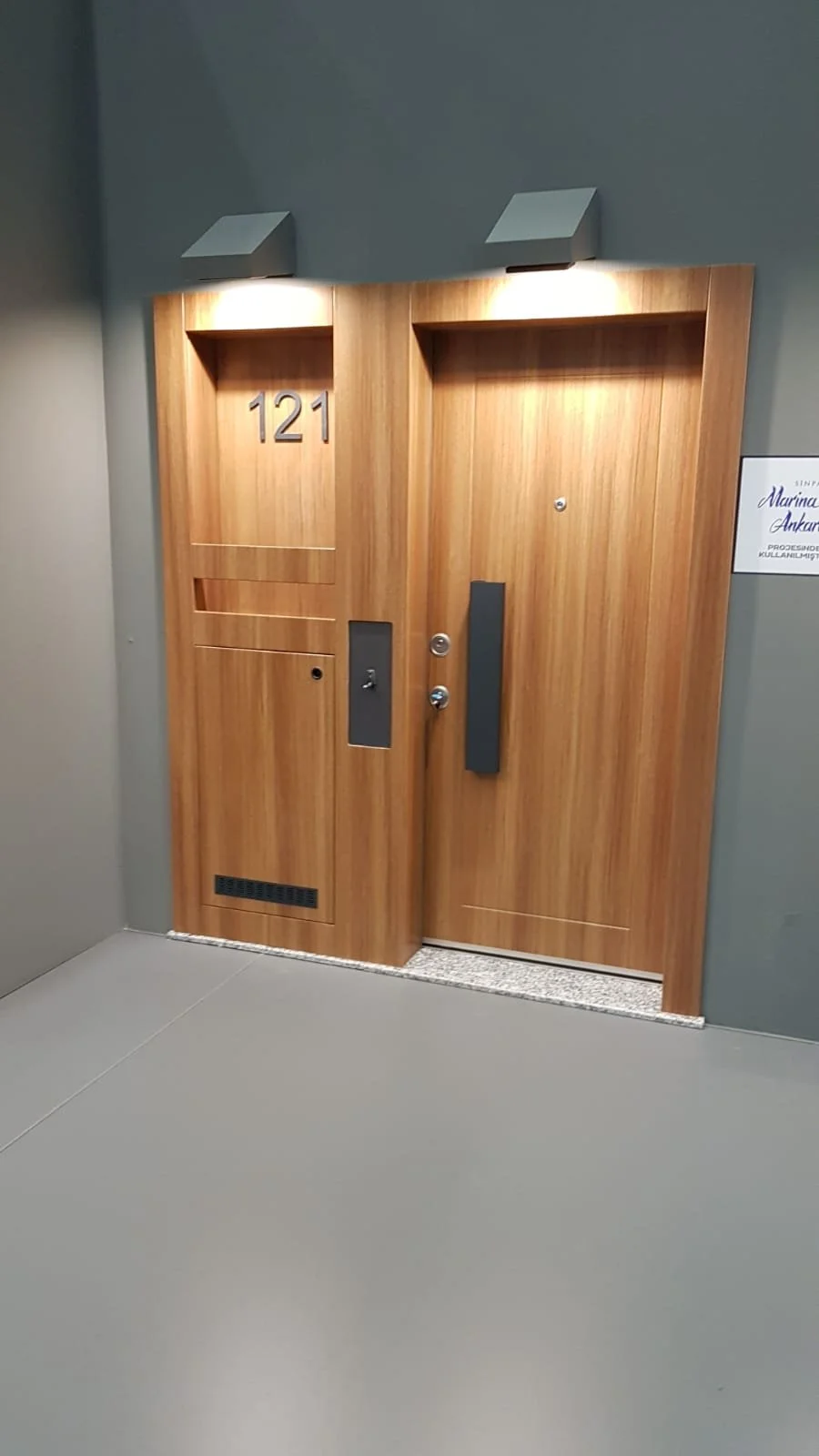 Wooden door with the number 121, featuring a mail slot, keyhole, and modern handle, illuminated by two overhead lights. Surrounding wall painted gray.