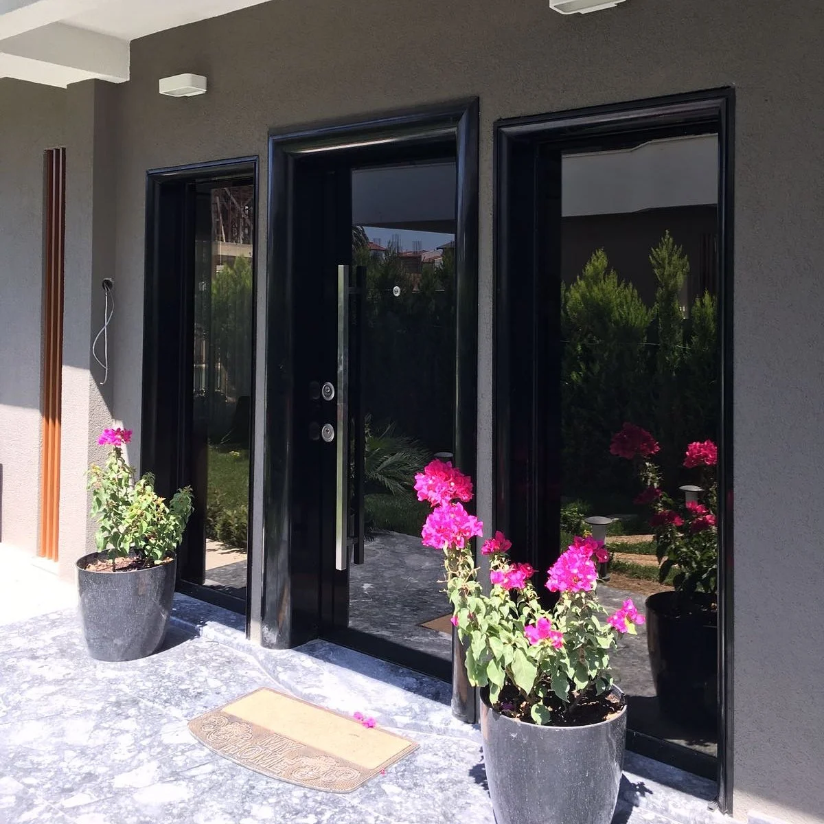 Modern front entrance with black glass door, pink flowering plants in black pots, and a welcome mat outside a gray house.