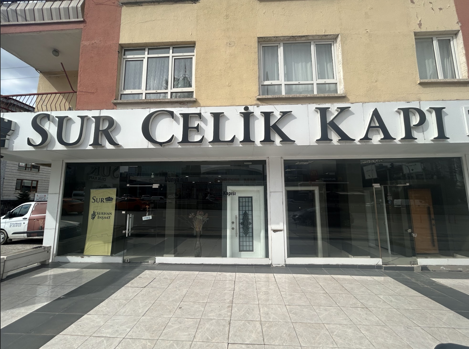 Ön cephesinde büyük siyah harflerle "SUR CELİK KAPI" yazan beyaz bir tabela, cam kapı ve pencereler ile dükkanın içinde küçük sarı bir tabela bulunan bir dükkân.