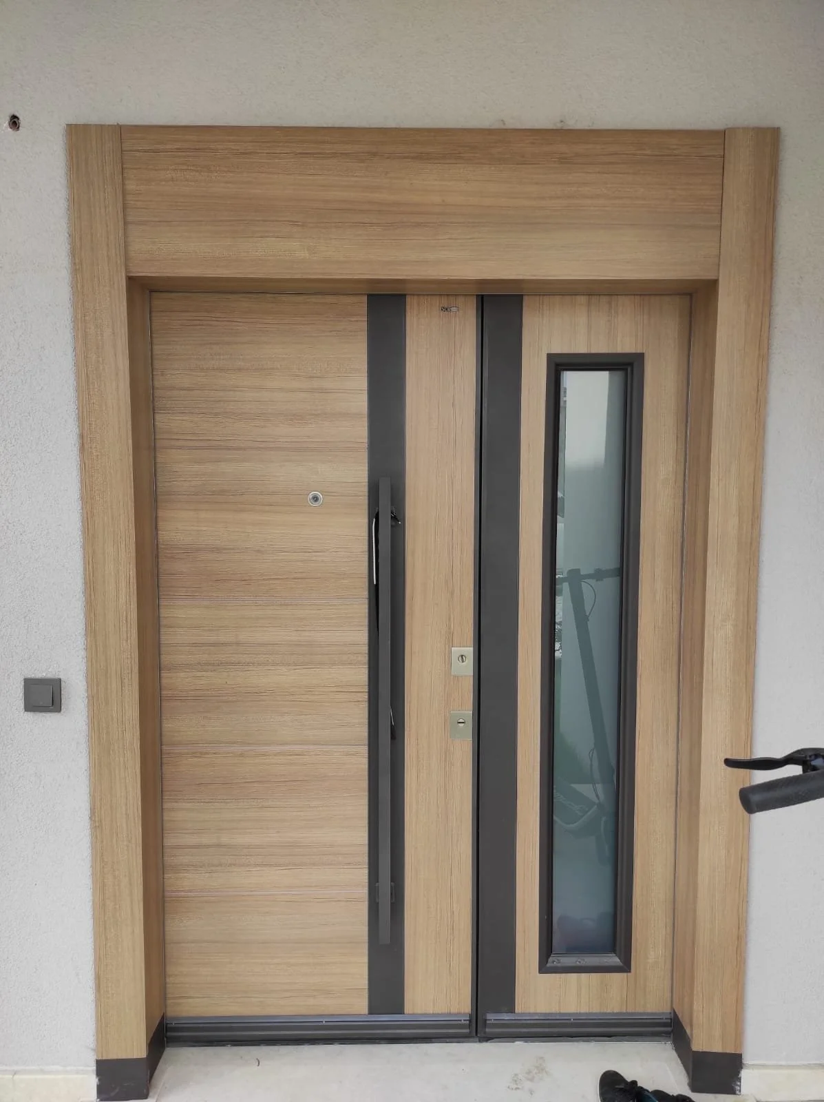 A modern wooden elevator door with a vertical narrow window and black frame, set in a white wall with a black corner trim, and a black bicycle is reflected in the window.