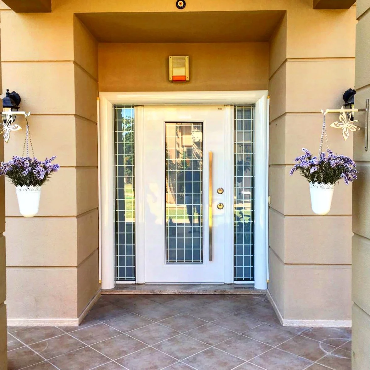 Front door of a house with decorative glass panels, flanked by two hanging flower pots with purple flowers, and a small porch area with tiled flooring.