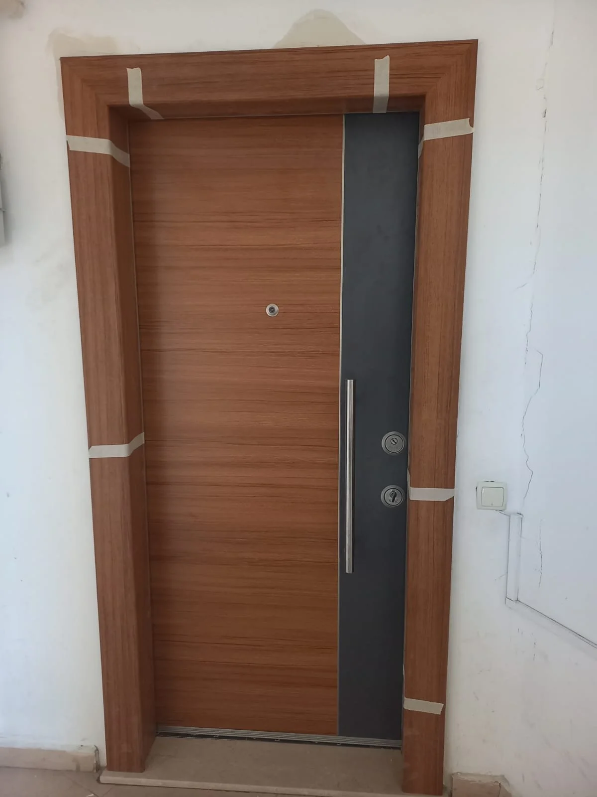 Wooden door with black vertical panel and long silver handle, surrounded by wooden trim, in a white wall with visible cracks and an electrical outlet.