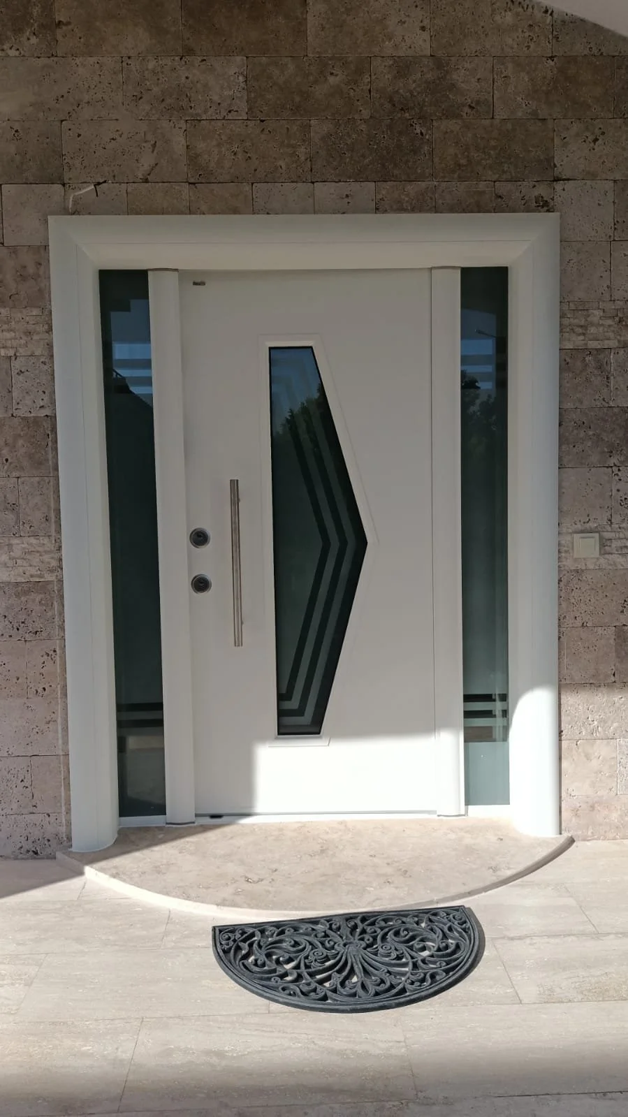 Geometrik cam panelleriyle süslenmiş, tuğla duvarla çevrili ve dekoratif siyah paspasla tamamlanmış modern beyaz ön kapı.