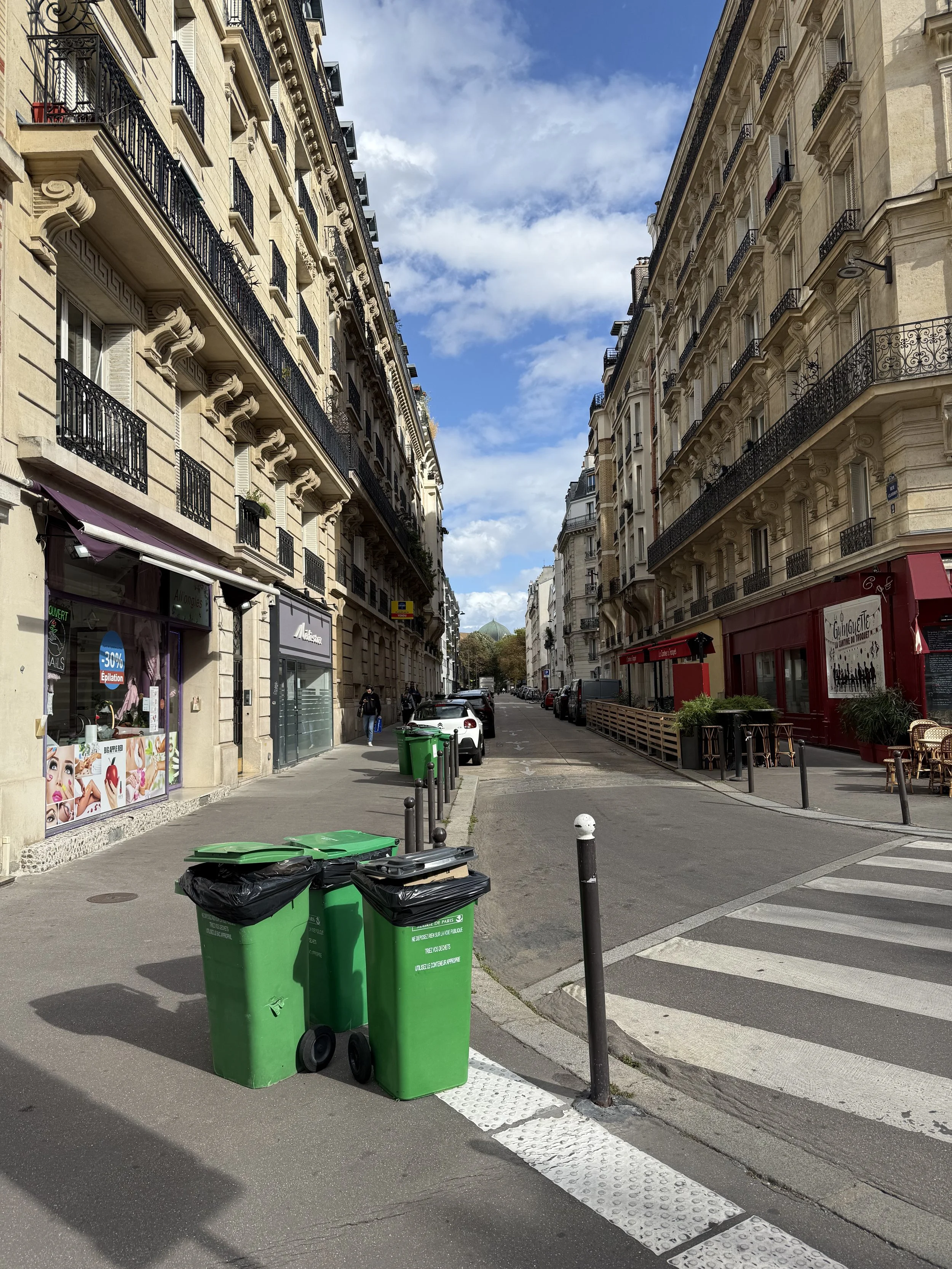 A quiet city street with multi-story beige buildings on both sides, with balconies and decorative facades. There are green and black trash bins and a few pedestrians, under a partly cloudy sky.
