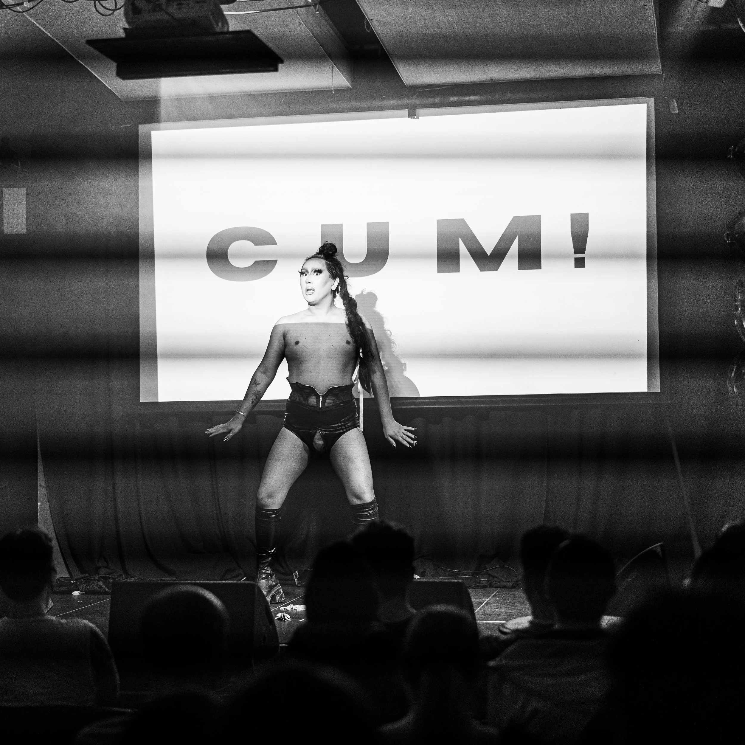 A black and white photograph of Dan Chan standing on stage in front of a projected image of the word "CUM!". They are performing an act for the cabaret night, SPEW. They are wearing a black corset, black briefs, and knee high black boots.