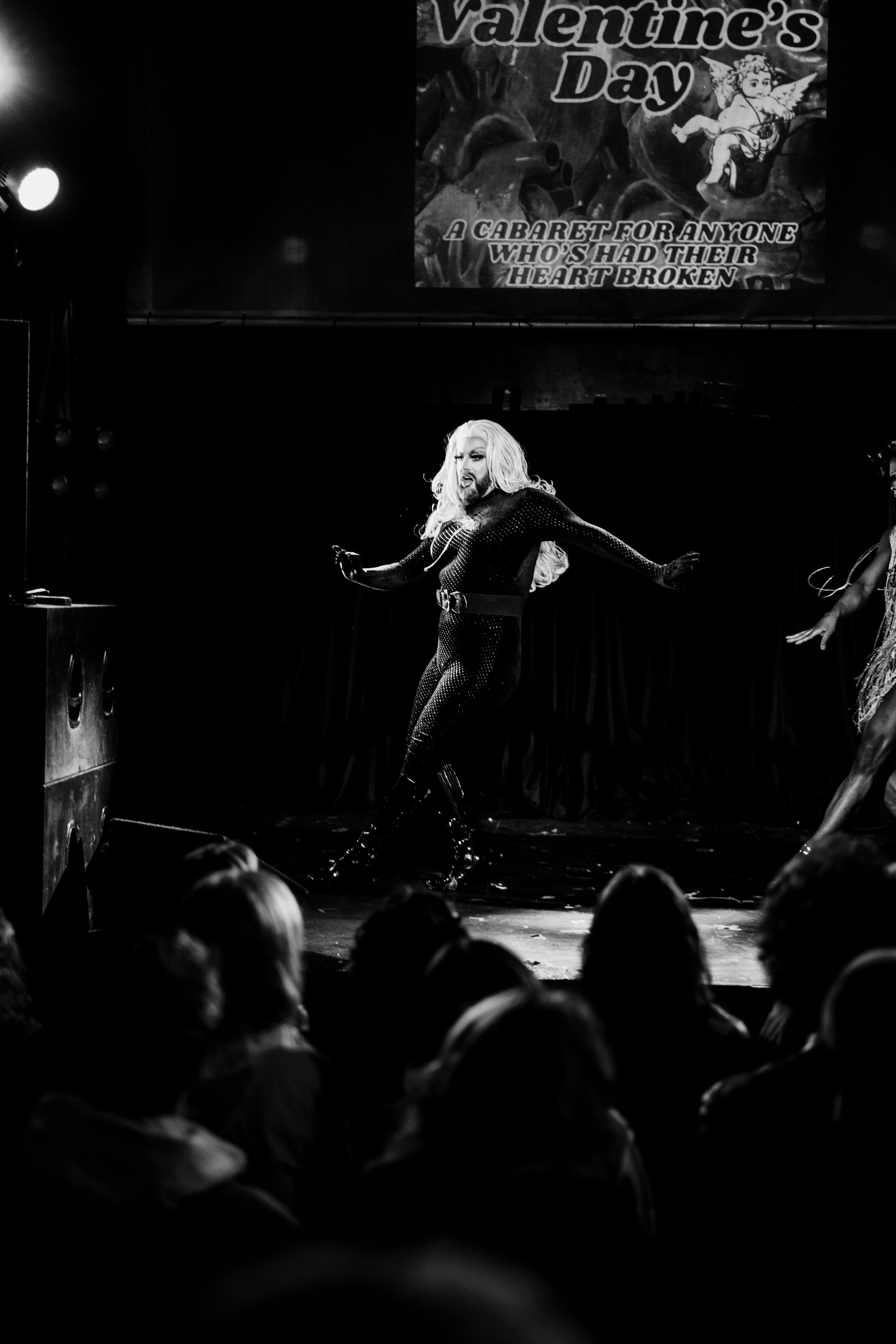 A performer on stage with long blonde hair, wearing a black outfit, performing at a Valentine's Day cabaret event. The stage background displays a sign reading 'Valentine's Day, A Cabaret for Anyone Who's Had Their Heart Broken,' with artwork of Cupid.