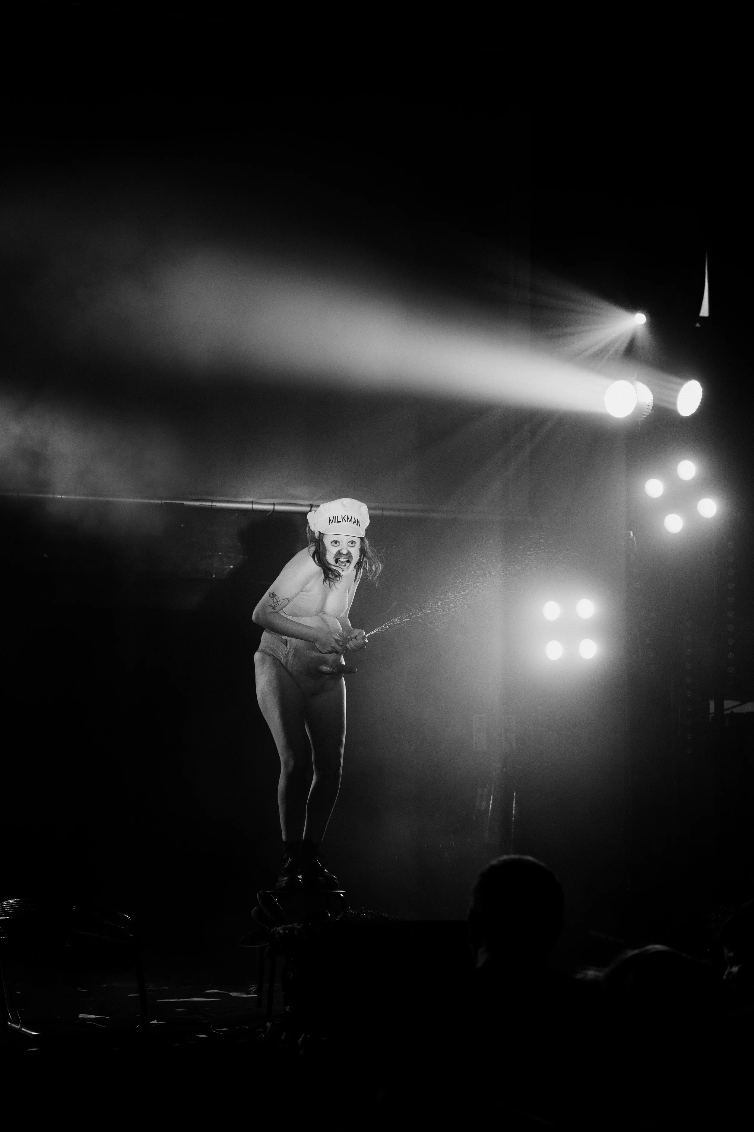 Black and white photo of a performer on stage wearing a oversized hat with 'Milkman' written on it, with stage lights shining in the background.